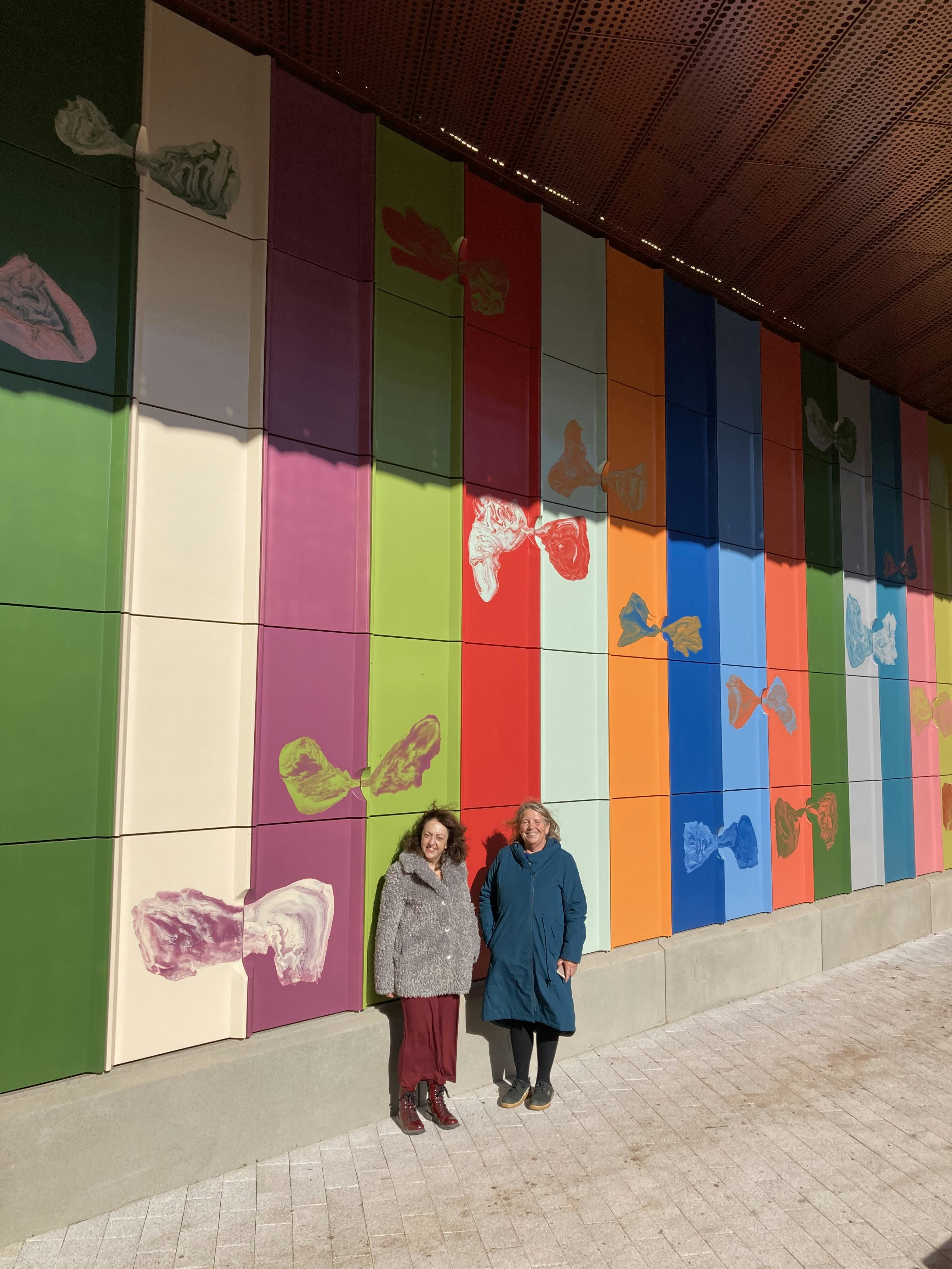 Two women stand in front of an artwork of large, colourful tiles