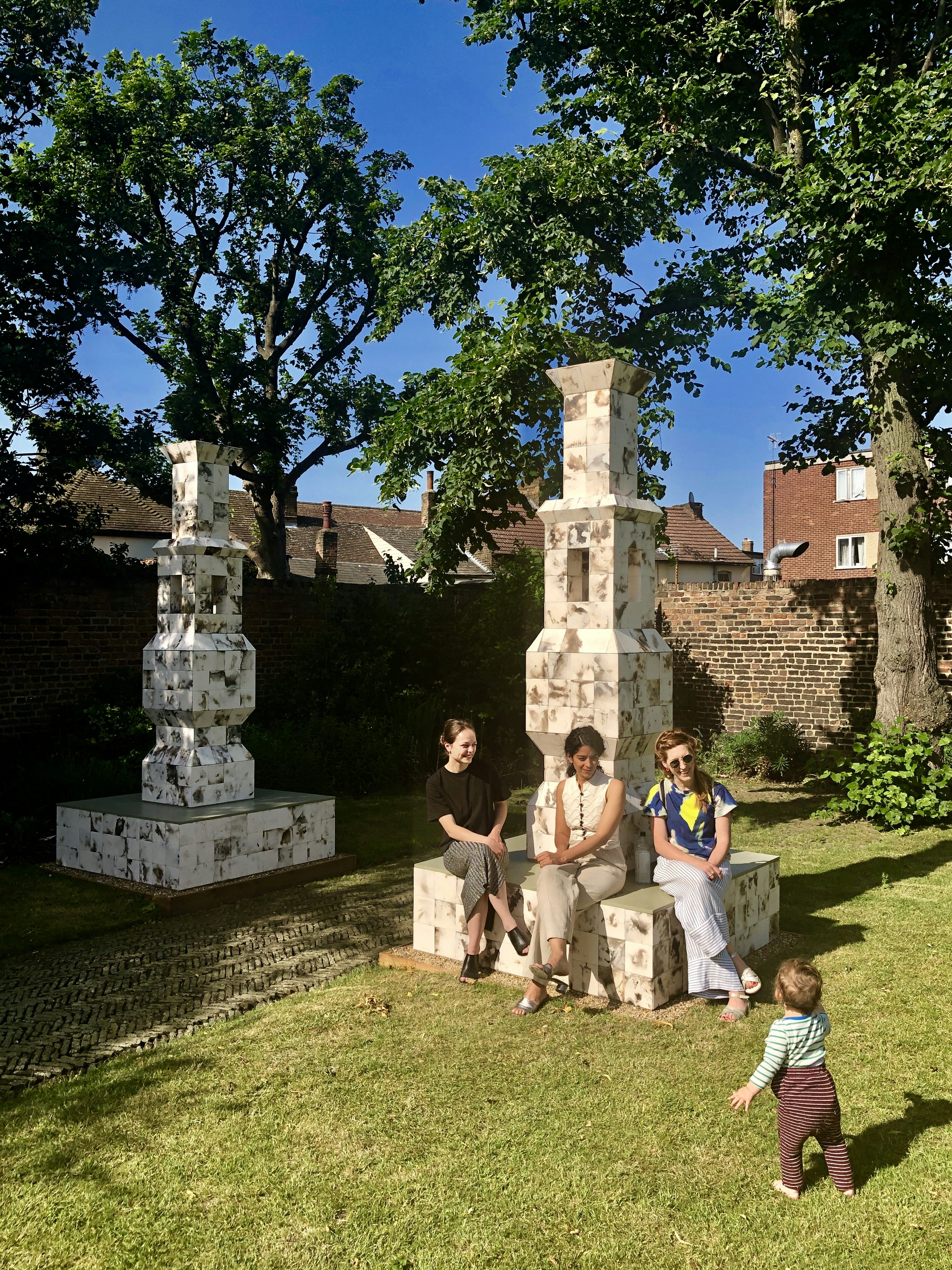 Three women sit on one of two tiled lanterns in a garden and look at a toddler in front of them