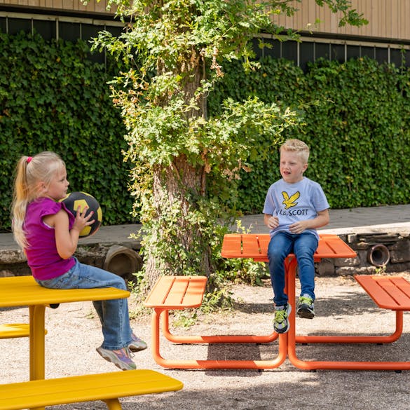 Spelende kinderen aan gekleurde metalen picknicktafels in zandbak school