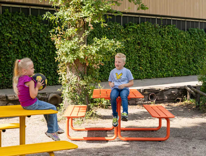 Spelende kinderen aan gekleurde metalen picknicktafels in zandbak school