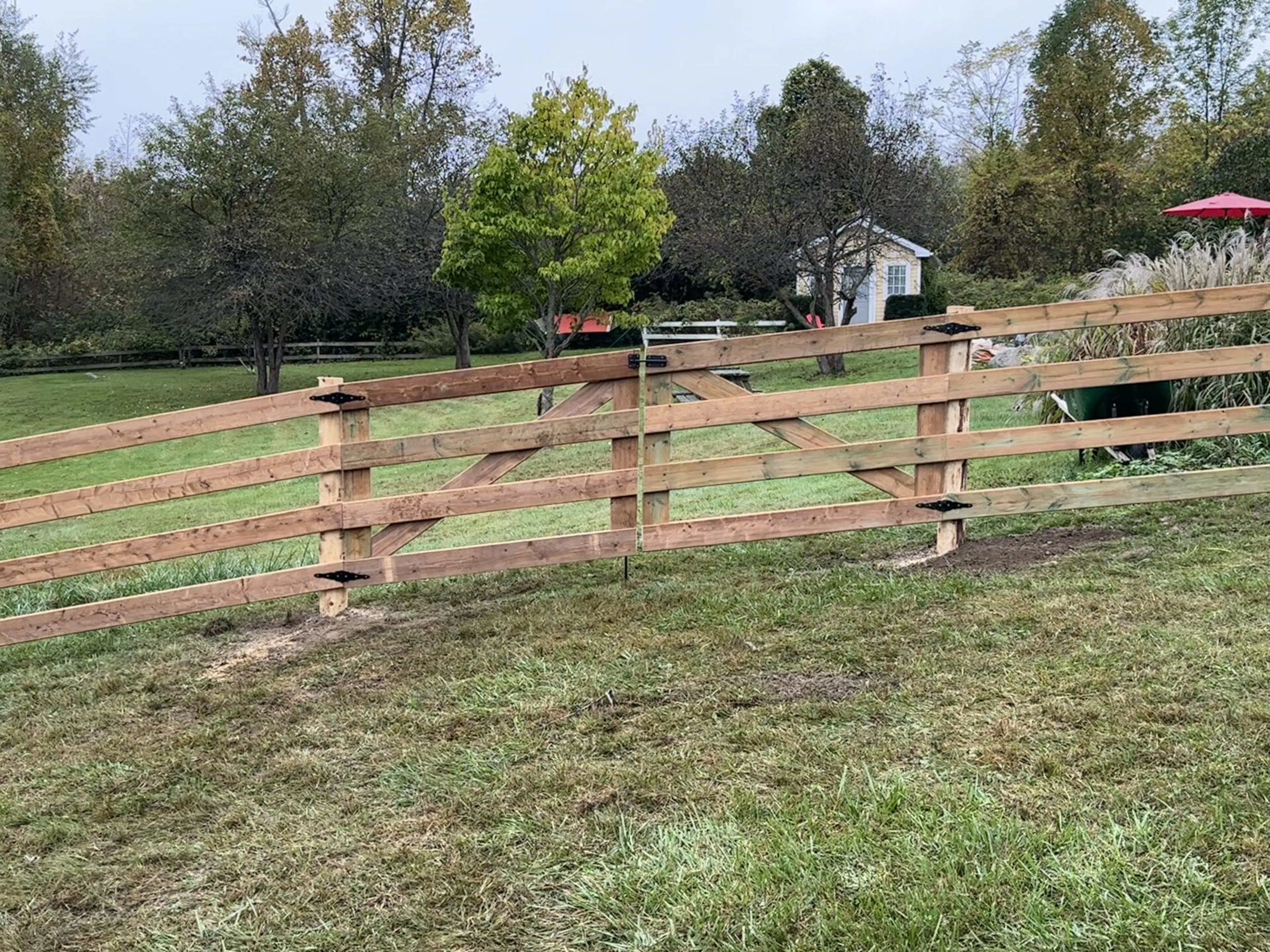 An agricultural boarded wooden fence with three horizontal rails is installed in a grassy yard with trees and a small building in the background.