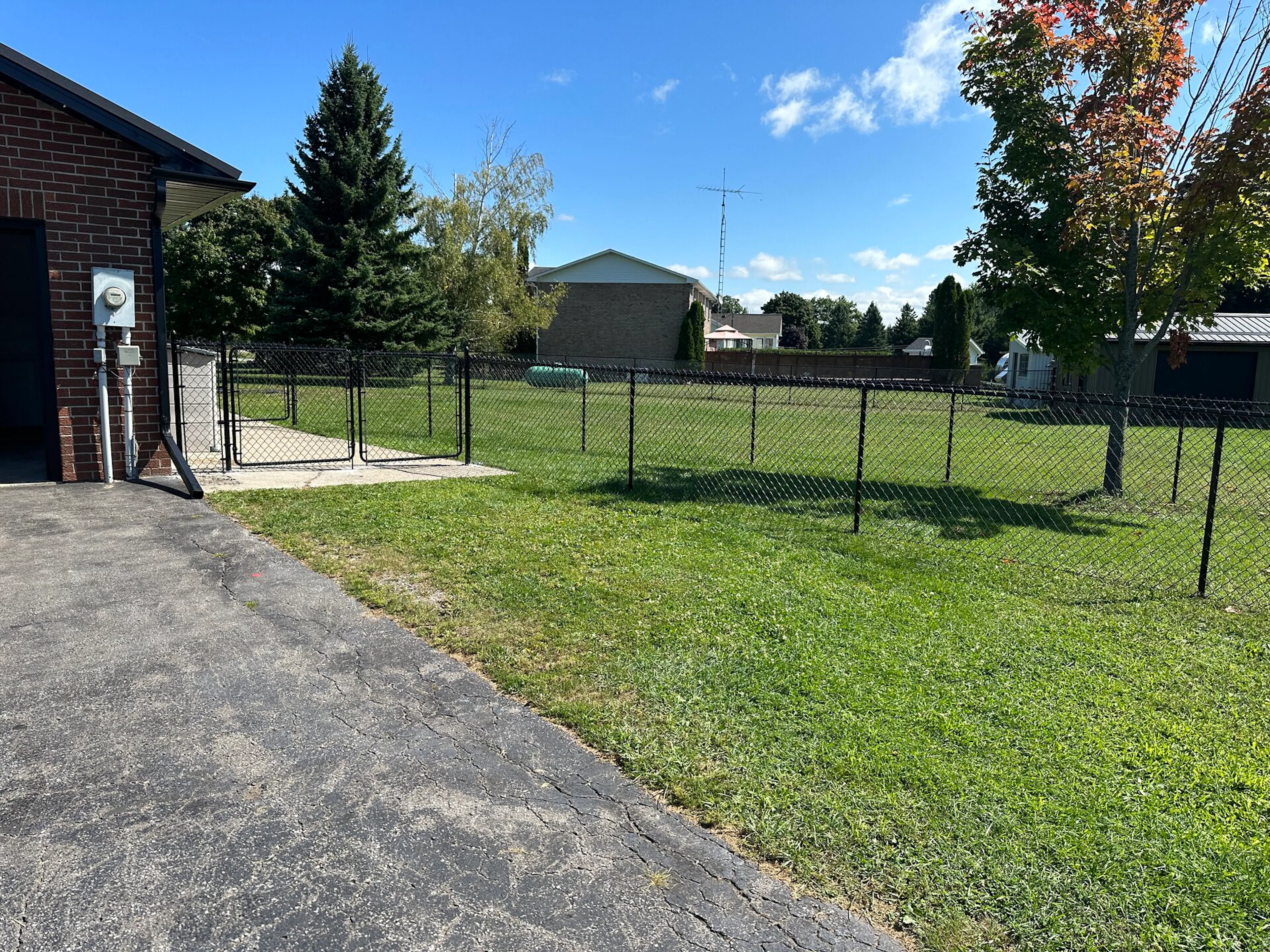 A fenced grassy area next to a brick building with trees and a clear blue sky in the background.