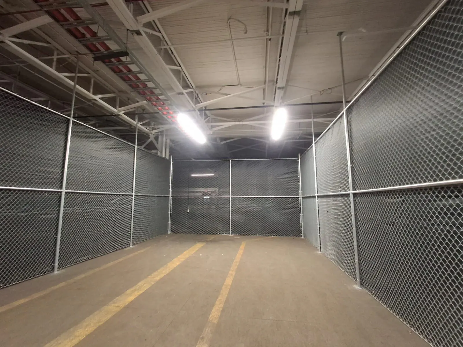 A view down a dimly lit commercial area with chain-link fences on both sides and fluorescent lights overhead.