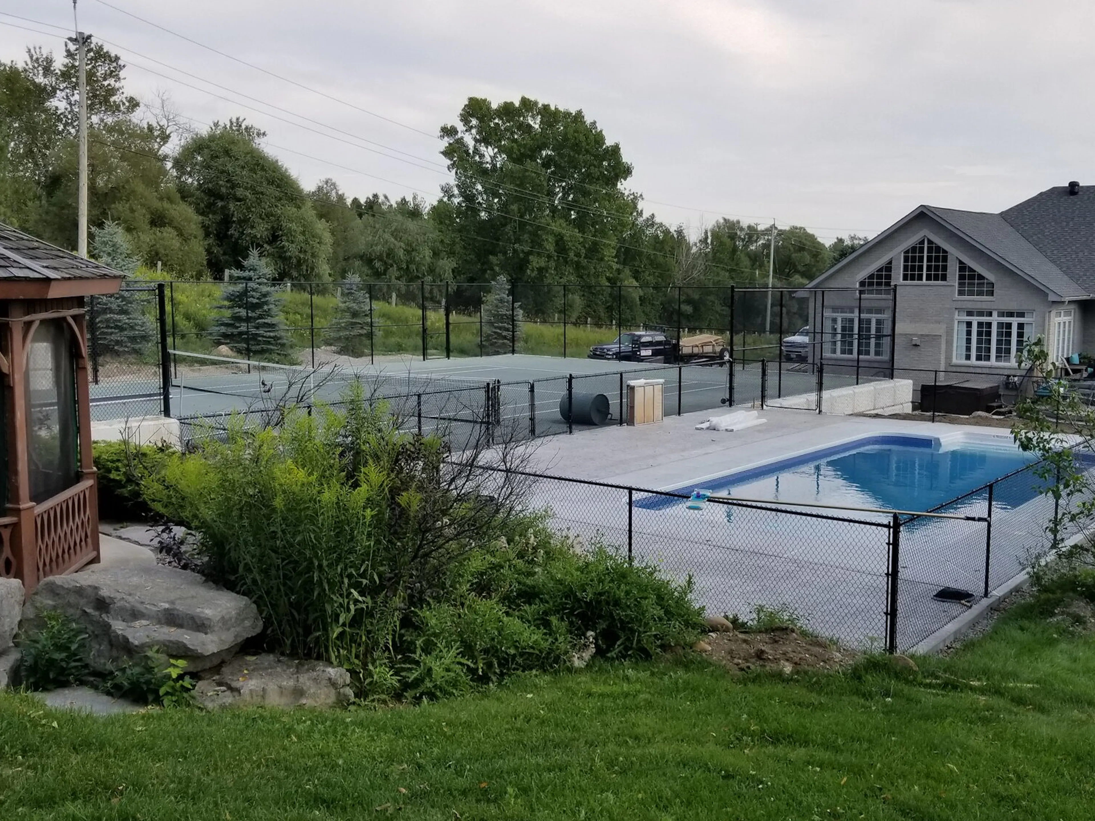 A swimming pool and tennis court enclosed at the perimeter of a residential backyard and throughout with a black chainlink fence.
