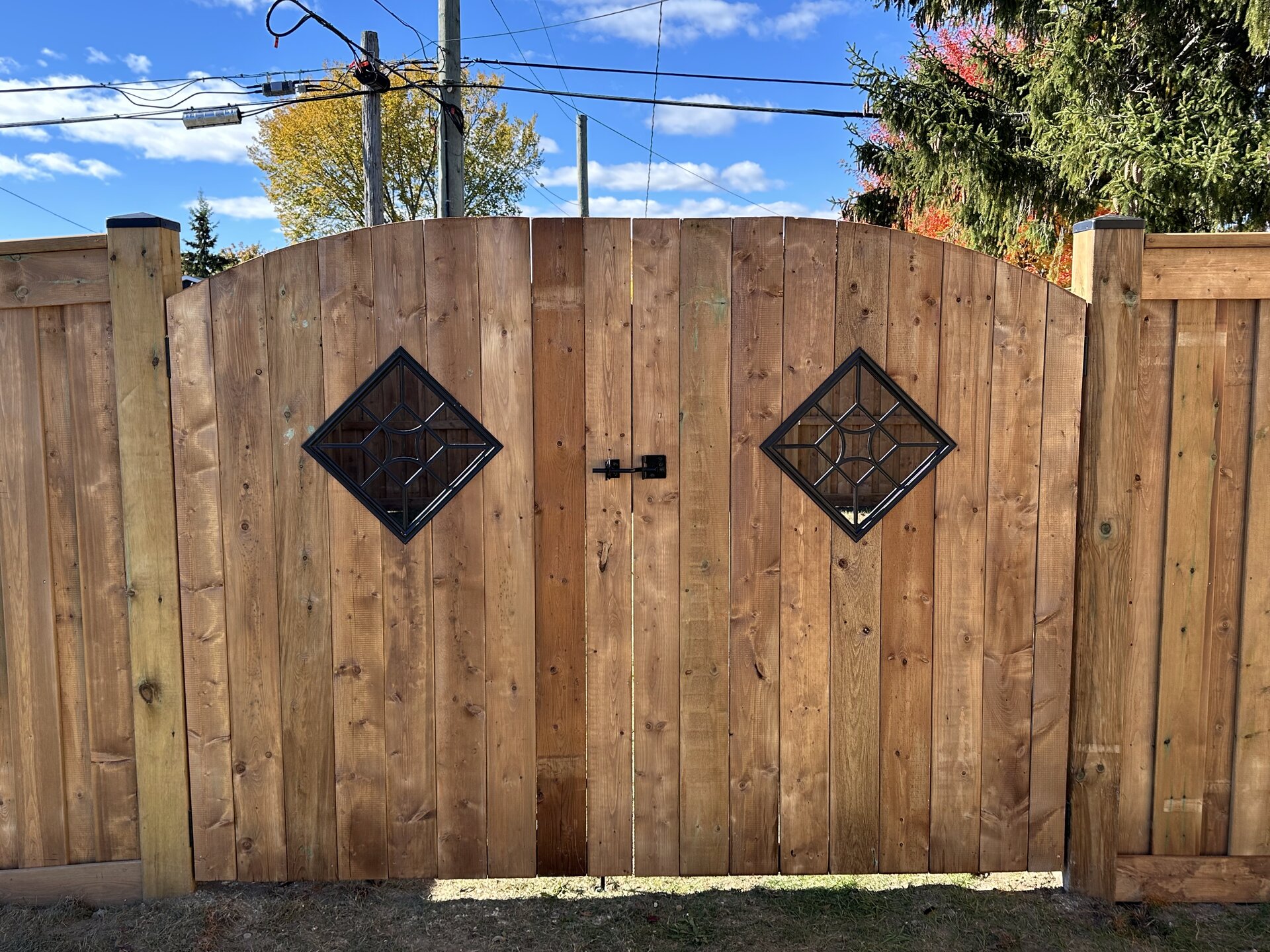 A wooden backyard gate with two diamond-shaped metal inserts and a black handle in the center of the fence gate.