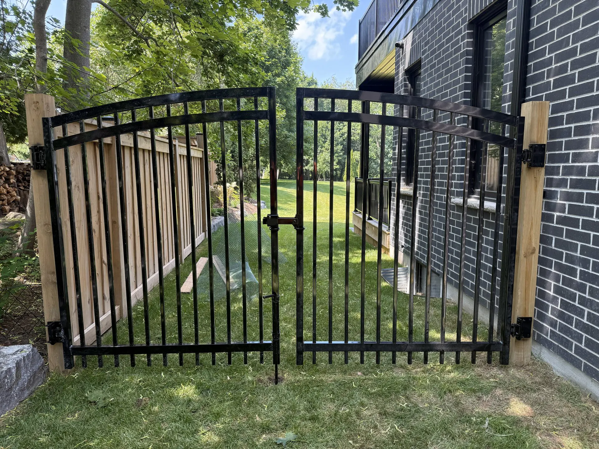 A black metal double ornamental gate with wooden posts stands in a grassy yard next to a brick building.