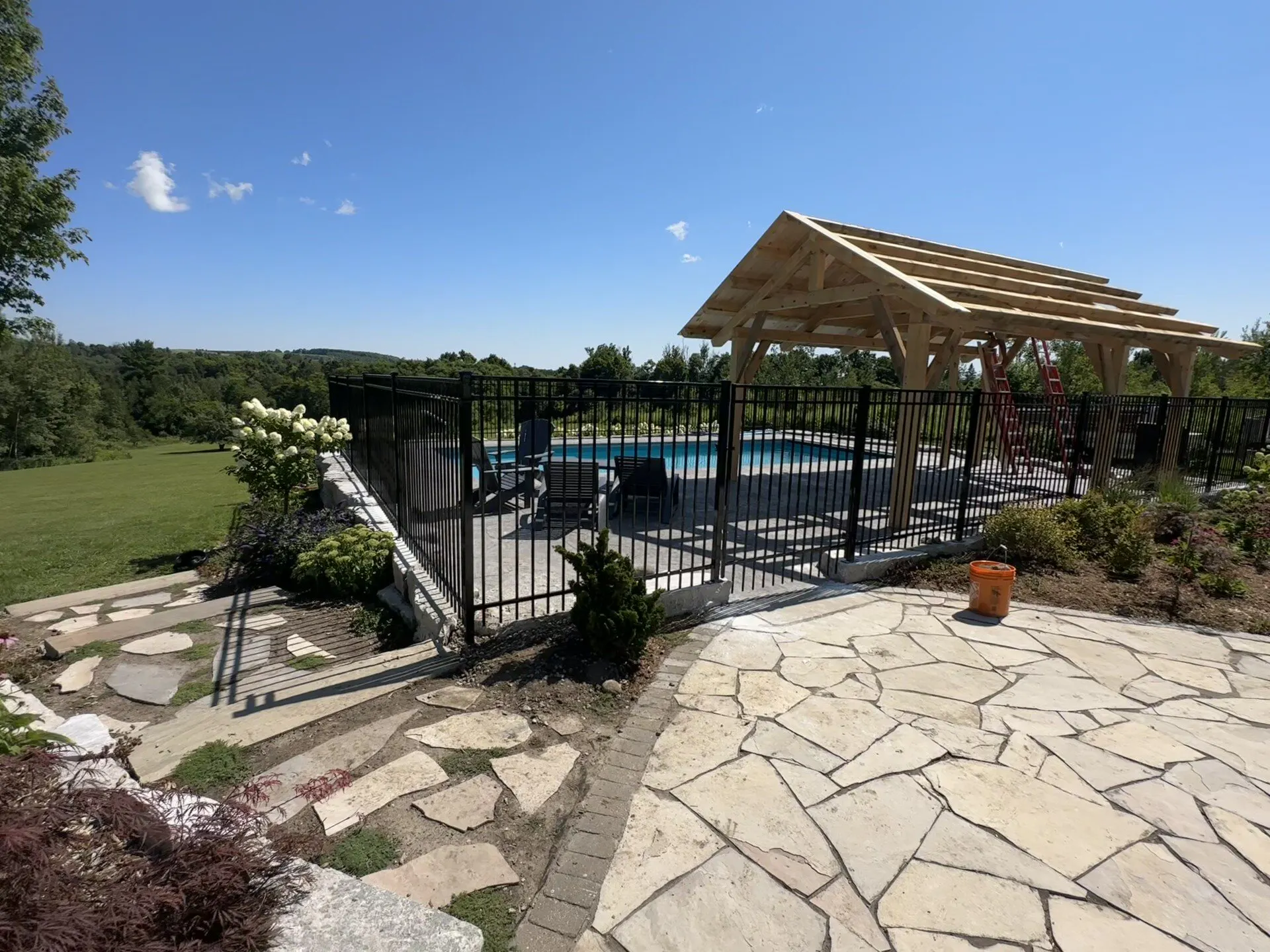 A backyard swimming pool area with a stone patio and a wooden pergola enclosed by a perimeter of black ornamental fence. Surrounded by lush greenery under a clear blue sky.