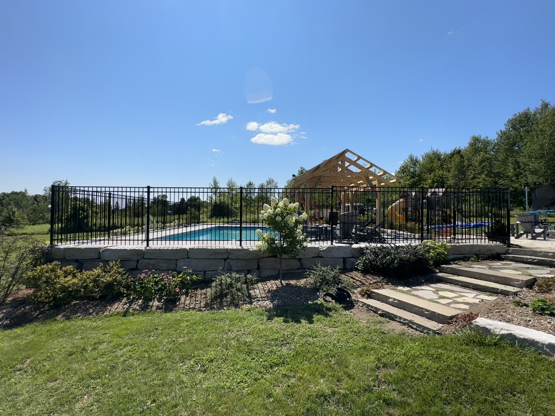 A backyard with a swimming pool enclosed by a black ornamental fence, with a gazebo, and a well-maintained lawn and garden area on a sunny day.