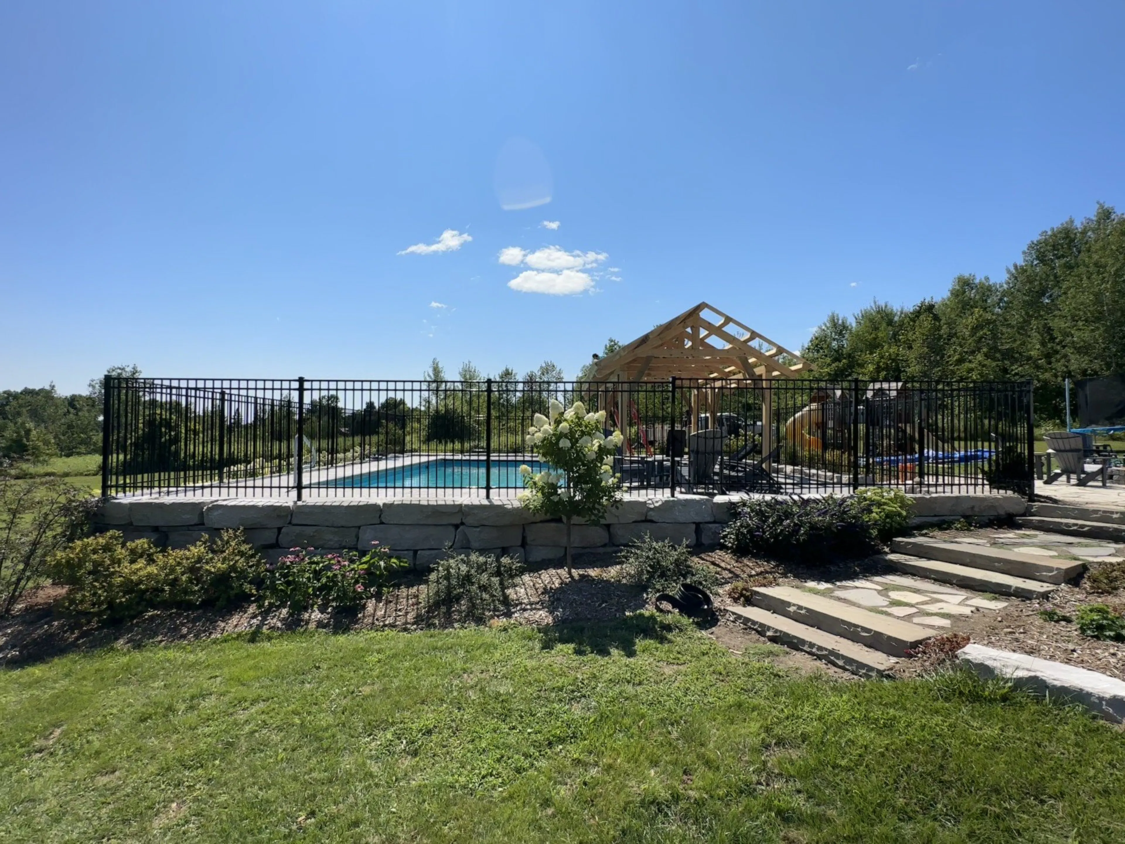 A backyard with a swimming pool enclosed by a black ornamental fence, with a gazebo, and a well-maintained lawn and garden area on a sunny day.