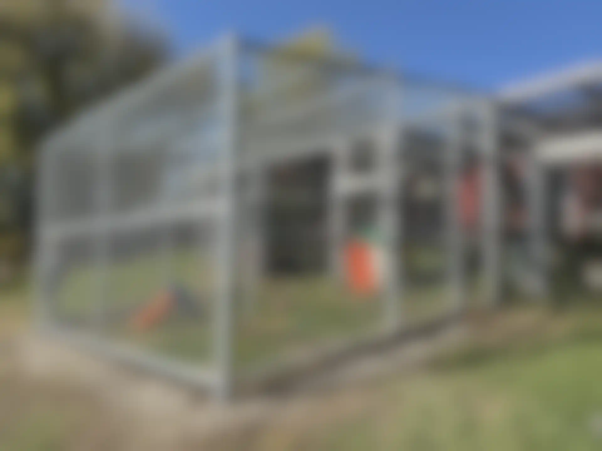 A large outdoor animal enclosure made of metal framing and wire mesh, with a small orange and white shelter inside and a slide on the left.