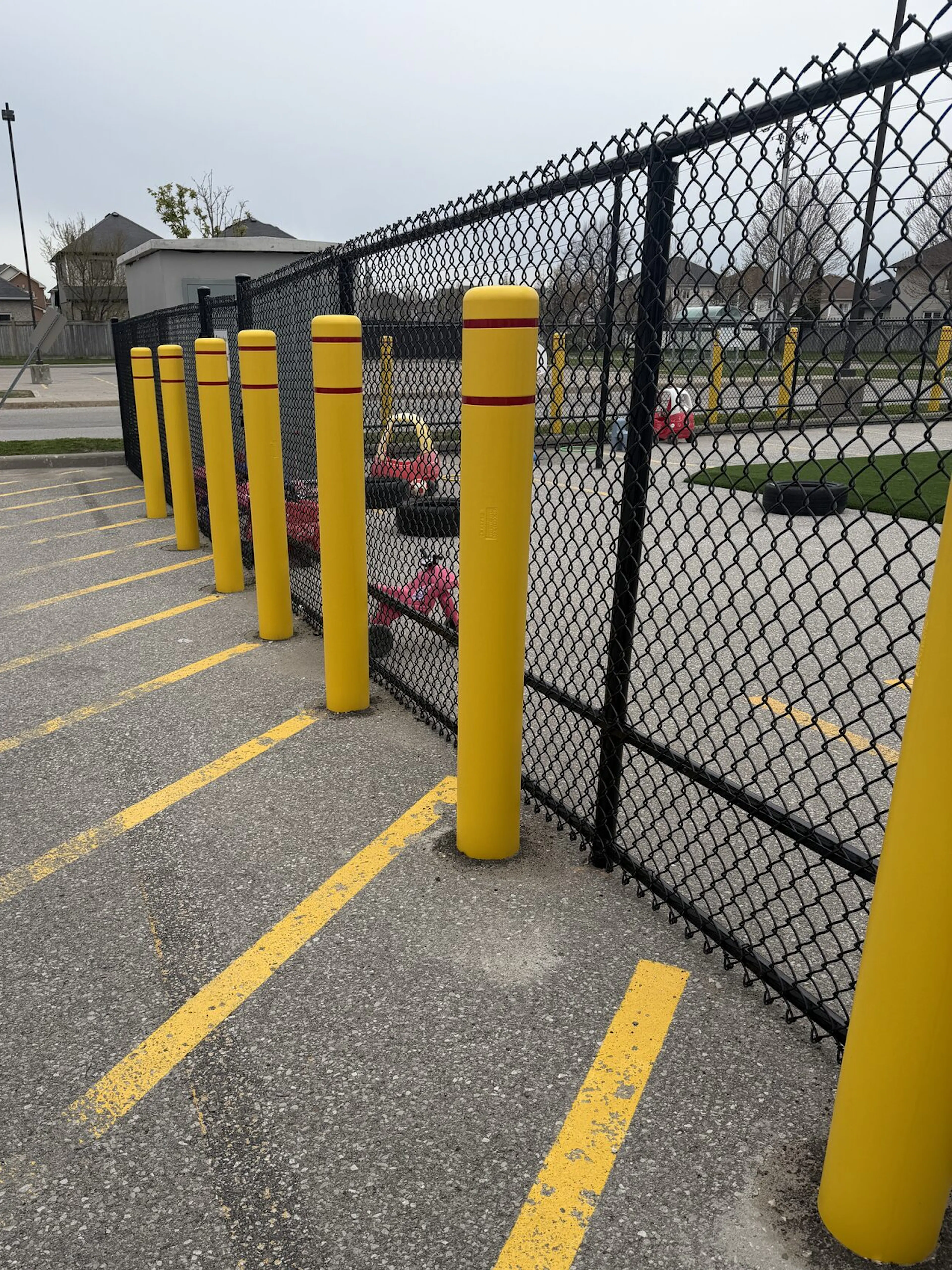 A sturdy black chain-link fence encloses a secure area, defined by bright yellow bollards that stand tall and straight at intervals along the perimeter. The asphalt surface beneath is marked with faded yellow lines, suggesting a parking lot. The overall design emphasizes safety and accessibility.
