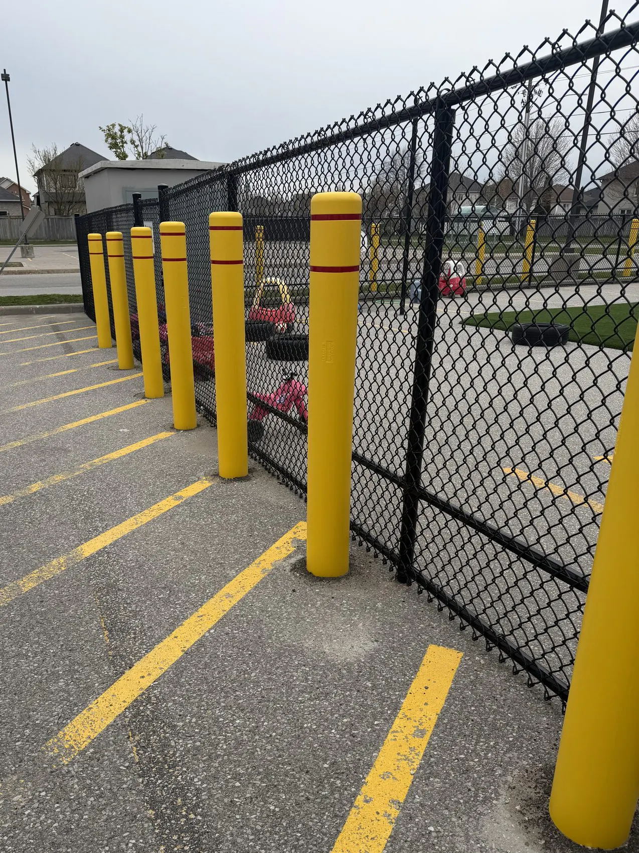 A sturdy black chain-link fence encloses a secure area, defined by bright yellow bollards that stand tall and straight at intervals along the perimeter. The asphalt surface beneath is marked with faded yellow lines, suggesting a parking lot. The overall design emphasizes safety and accessibility.