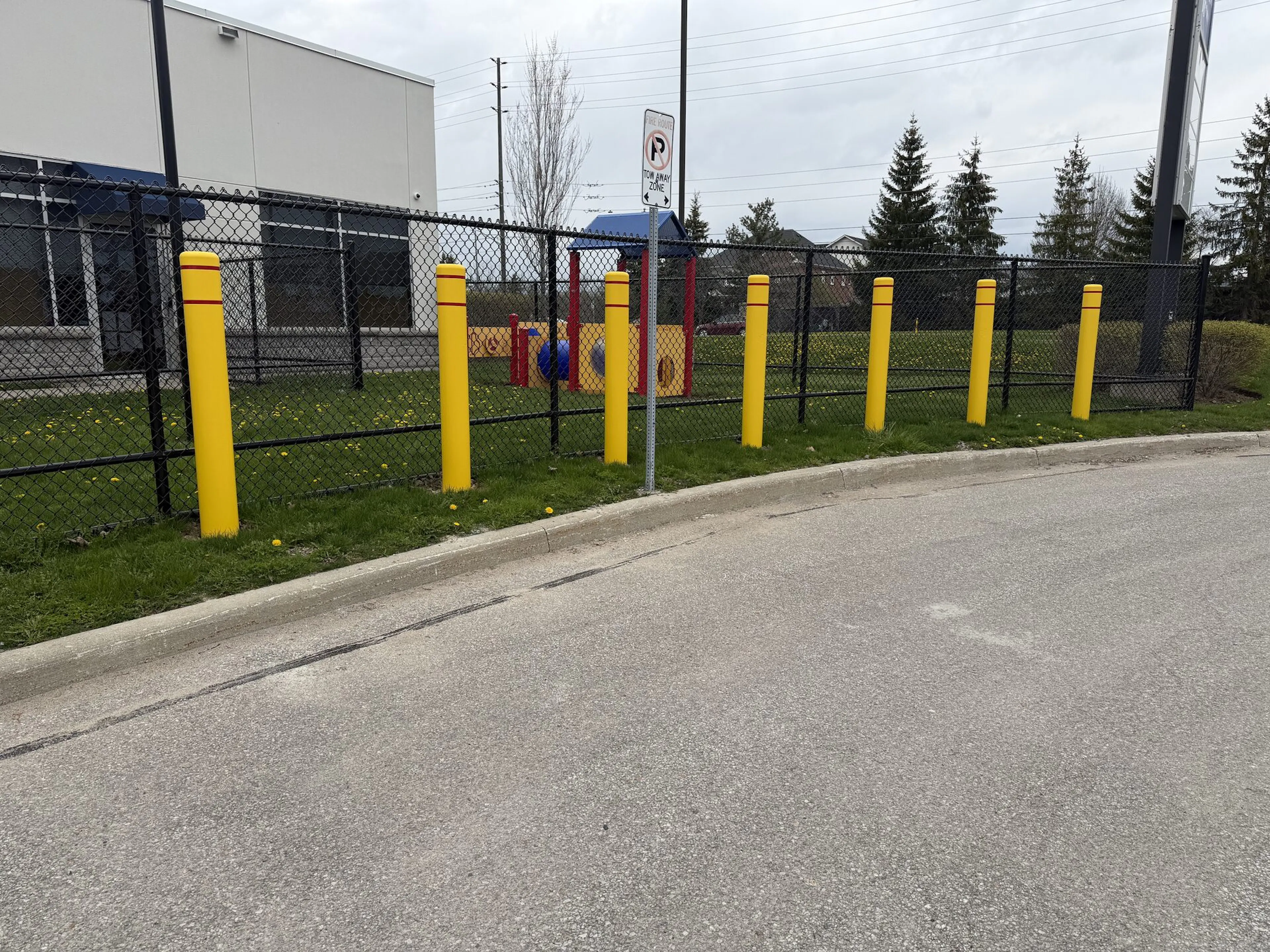 A sturdy black security fence enclosing a grassy perimeter, accented by bright yellow posts that provide additional protection. The posts, arranged at regular intervals, contrast sharply against the muted tones of a cloudy sky and an adjacent gray pavement.