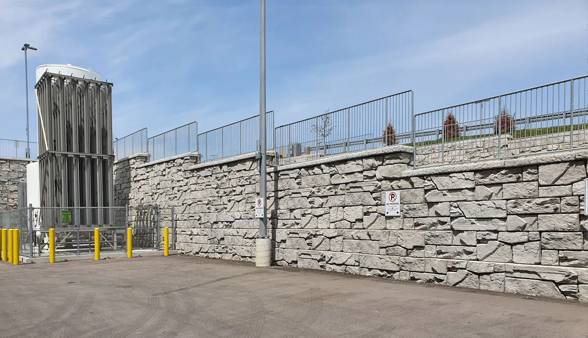A robust stone wall serves as the perimeter, topped with a low metal fence that enhances security. The structure is complemented by a towering guard tower, casting shadows over a sunlit, paved area. Clear blue skies create a stark contrast against the gray stone, emphasizing the imposing nature of the gates.
