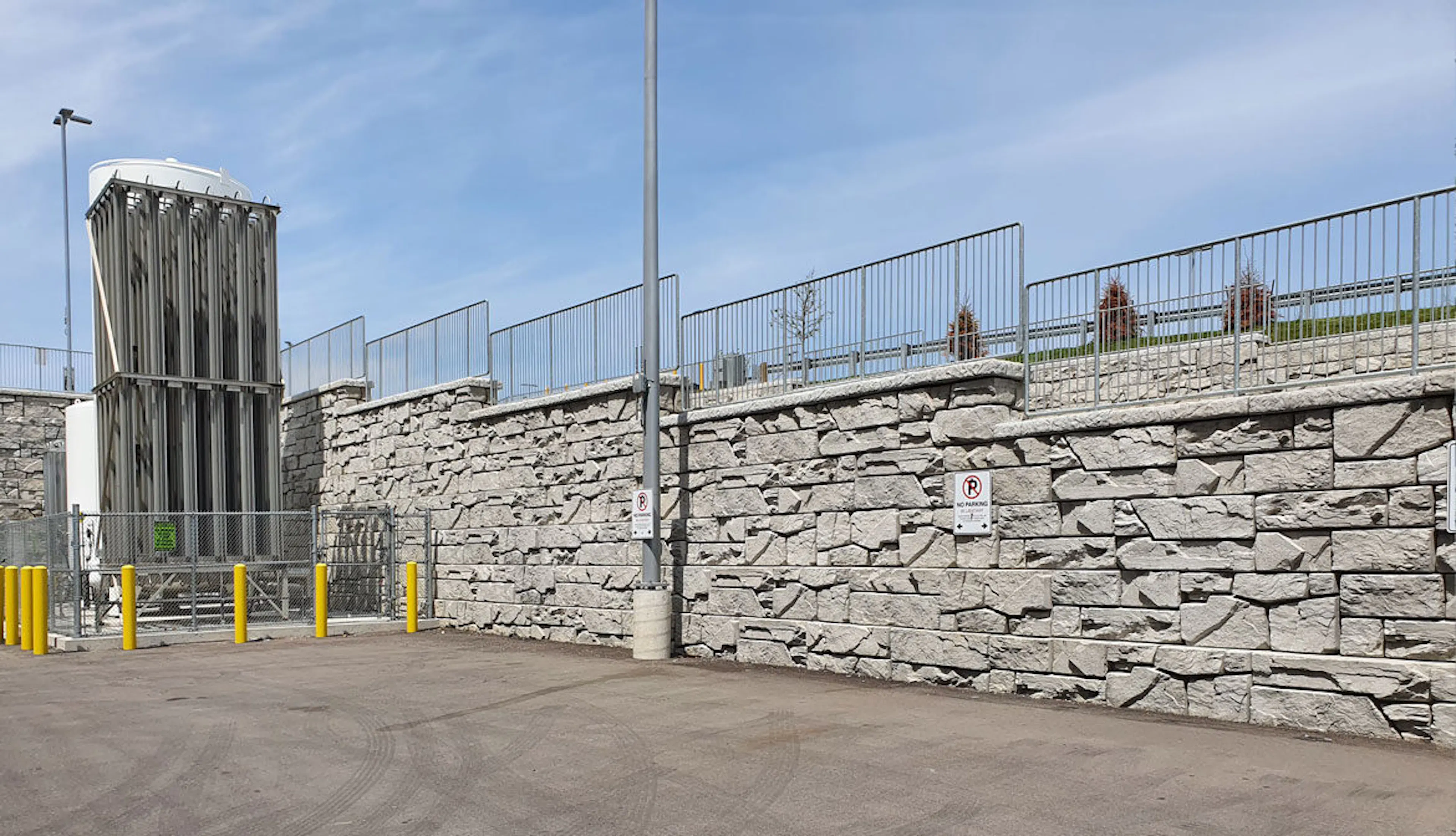 A robust stone wall serves as the perimeter, topped with a low metal fence that enhances security. The structure is complemented by a towering guard tower, casting shadows over a sunlit, paved area. Clear blue skies create a stark contrast against the gray stone, emphasizing the imposing nature of the gates.