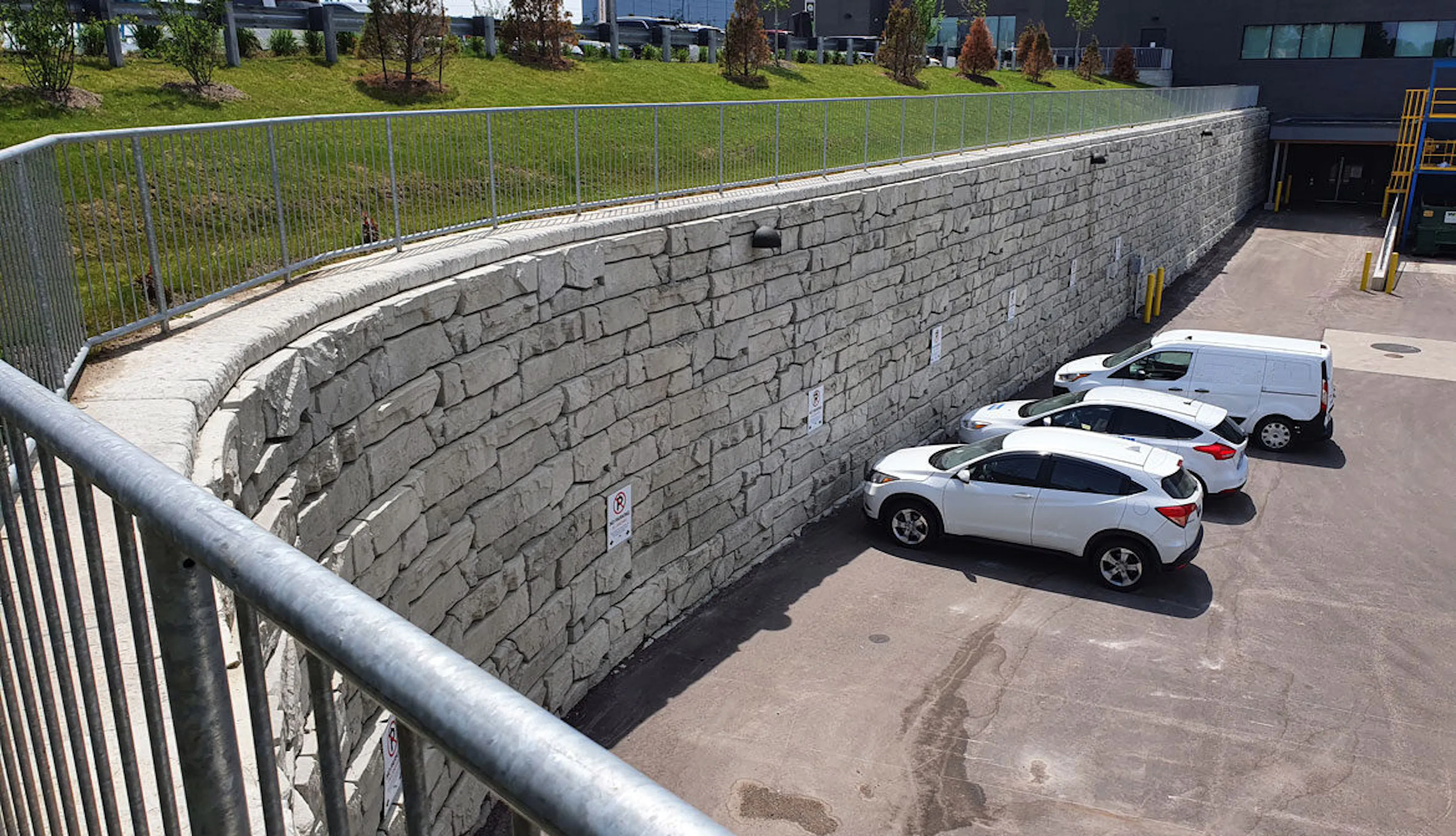 A view from a raised vantage point of a modern parking area enclosed by a sturdy stone wall, forming a secure perimeter. Two sleek white vehicles are parked closely together on the open lot, with a green lawn and buildings visible in the distance, suggesting a clear boundary defined by gates.