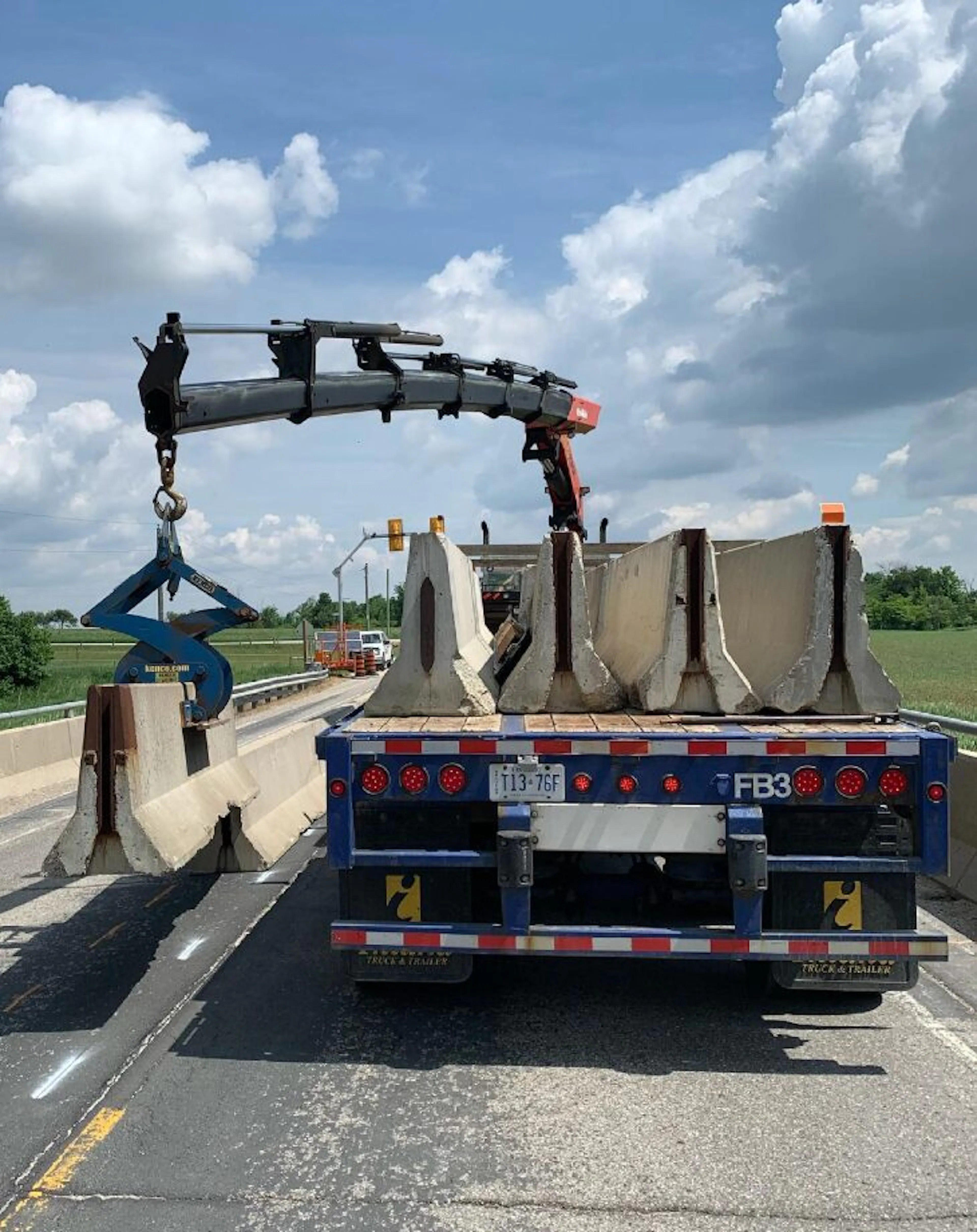 A large flatbed truck on a highway features a crane extending over the top, preparing to lift concrete barriers for construction. The sky is partly cloudy, casting soft shadows on the green landscape, emphasizing the truck’s role in enhancing security along the perimeter.