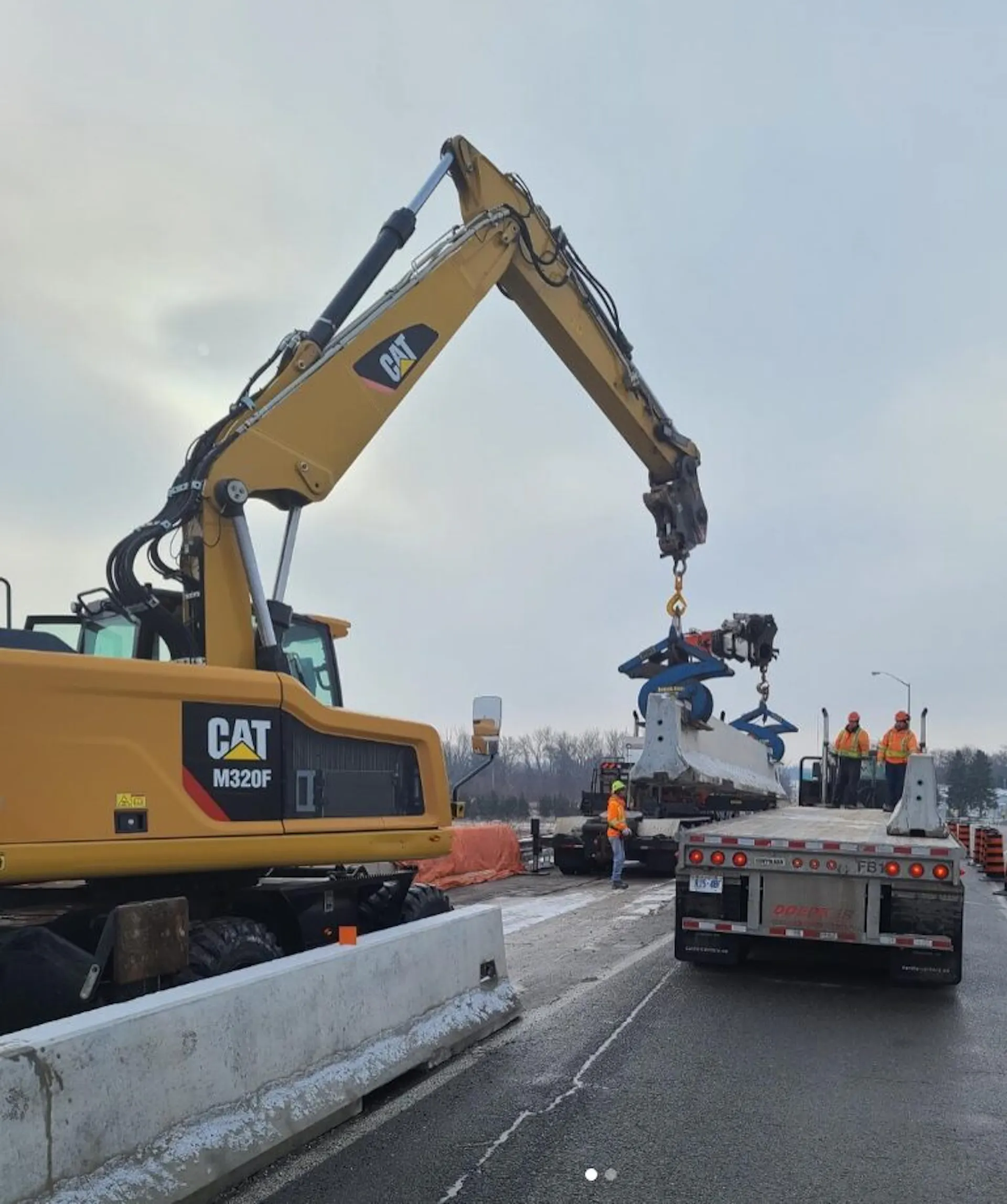 A robust yellow excavator, branded with the CAT logo, maneuvers laboriously above a construction site. Its long arm extends gracefully, wielding a large metal claw poised to pick up debris. In the background, a concrete barrier separates ongoing work from vehicular traffic, enhancing the perimeter security of the area.