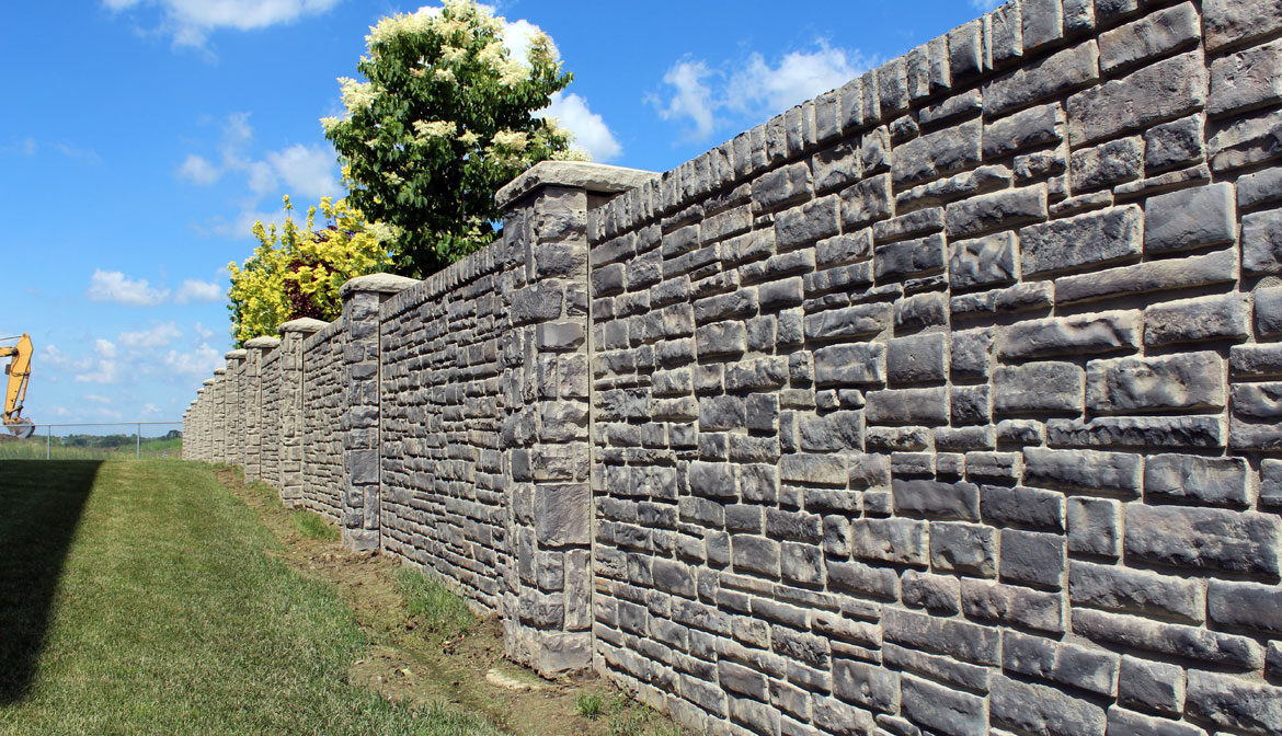 A grey concrete stone fence with bushes and trees behind it under a blue sky with clouds.