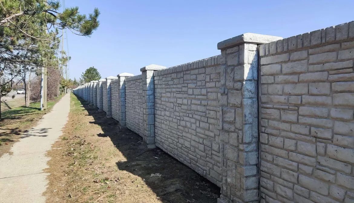 A long, light gray stone wall with concrete posts runs alongside a paved walkway under a clear blue sky.