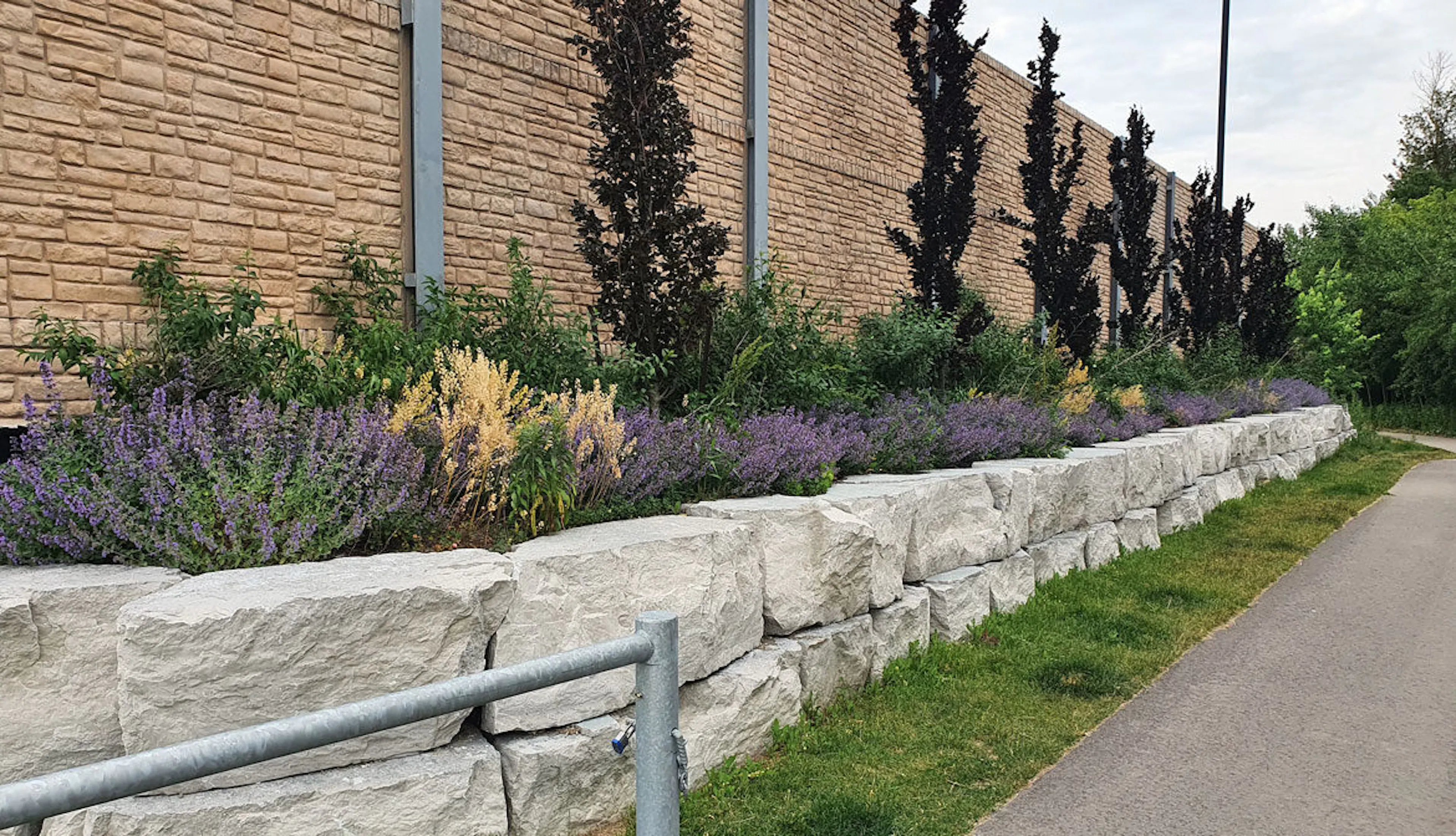 A grey concrete stone retaining wall with purple and yellow flowers and dark trees behind it.