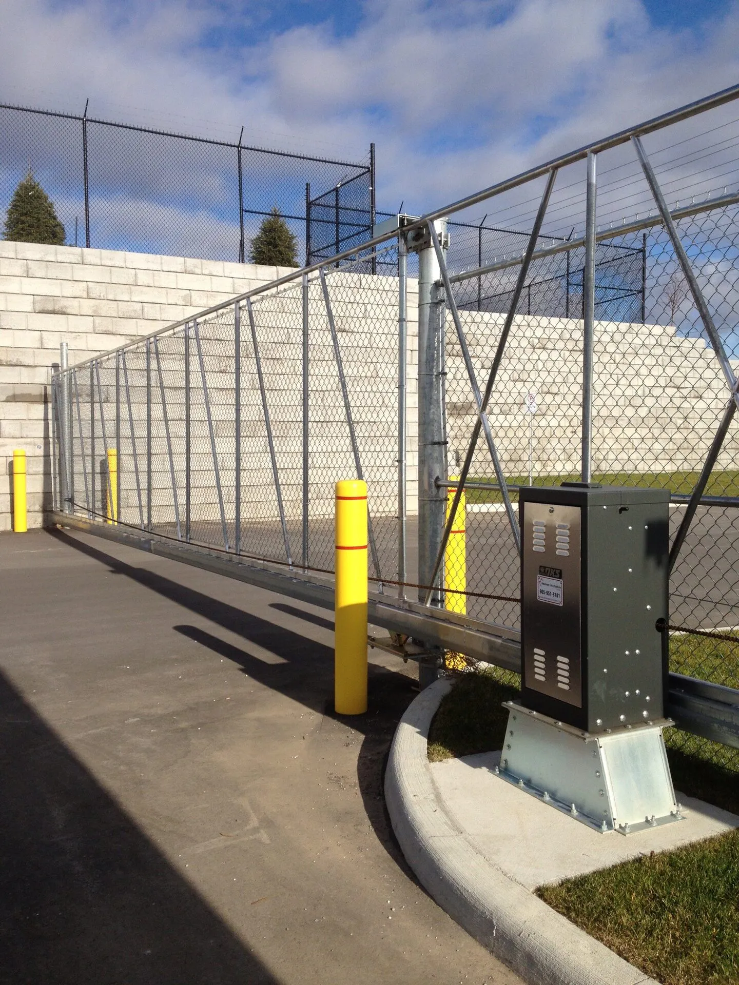 A slide chain-link fence gate with yellow posts and a control panel, leading to a parking area with a stone wall in the background.