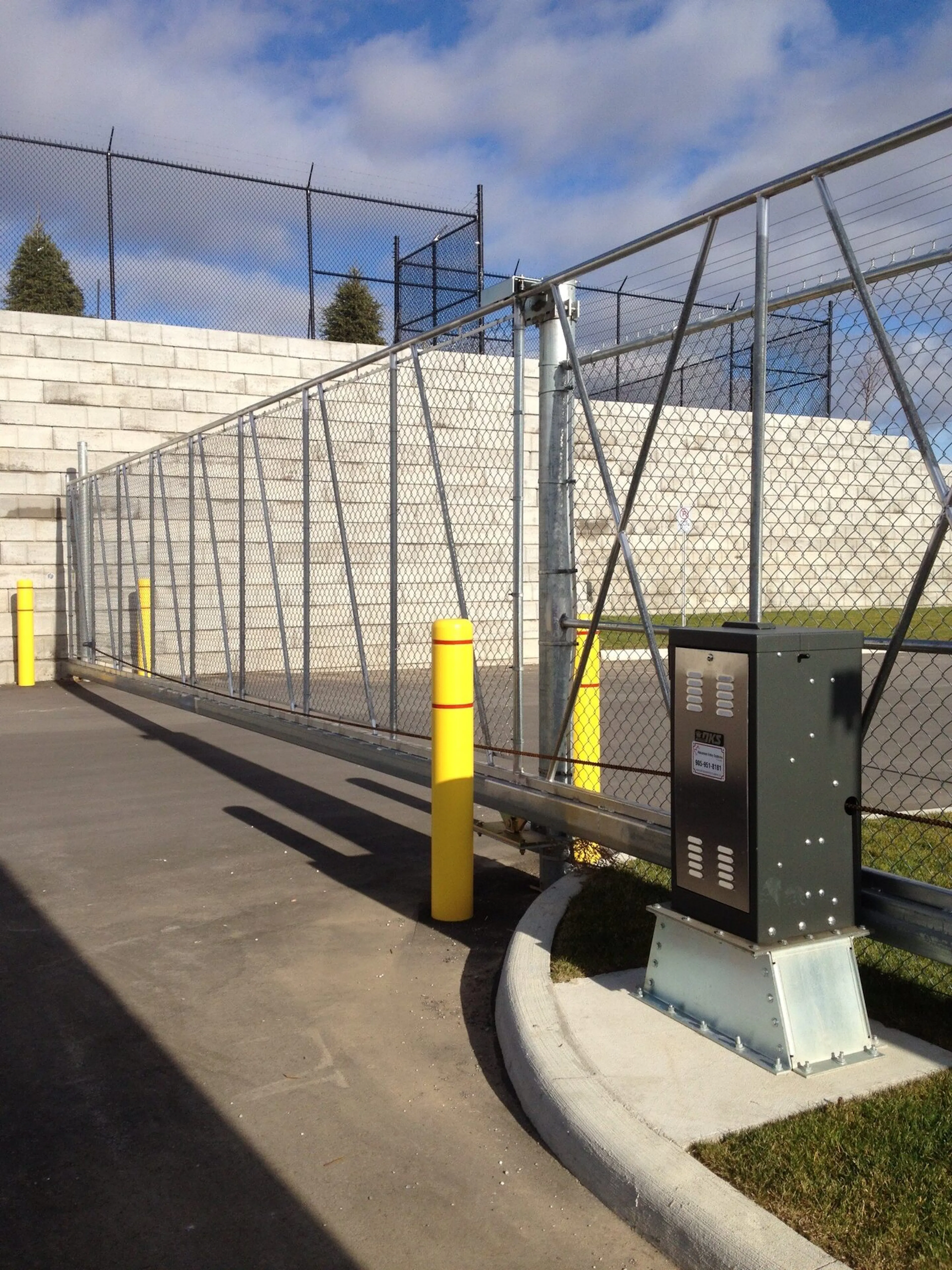 A slide chain-link fence gate with yellow posts and a control panel, leading to a parking area with a stone wall in the background.