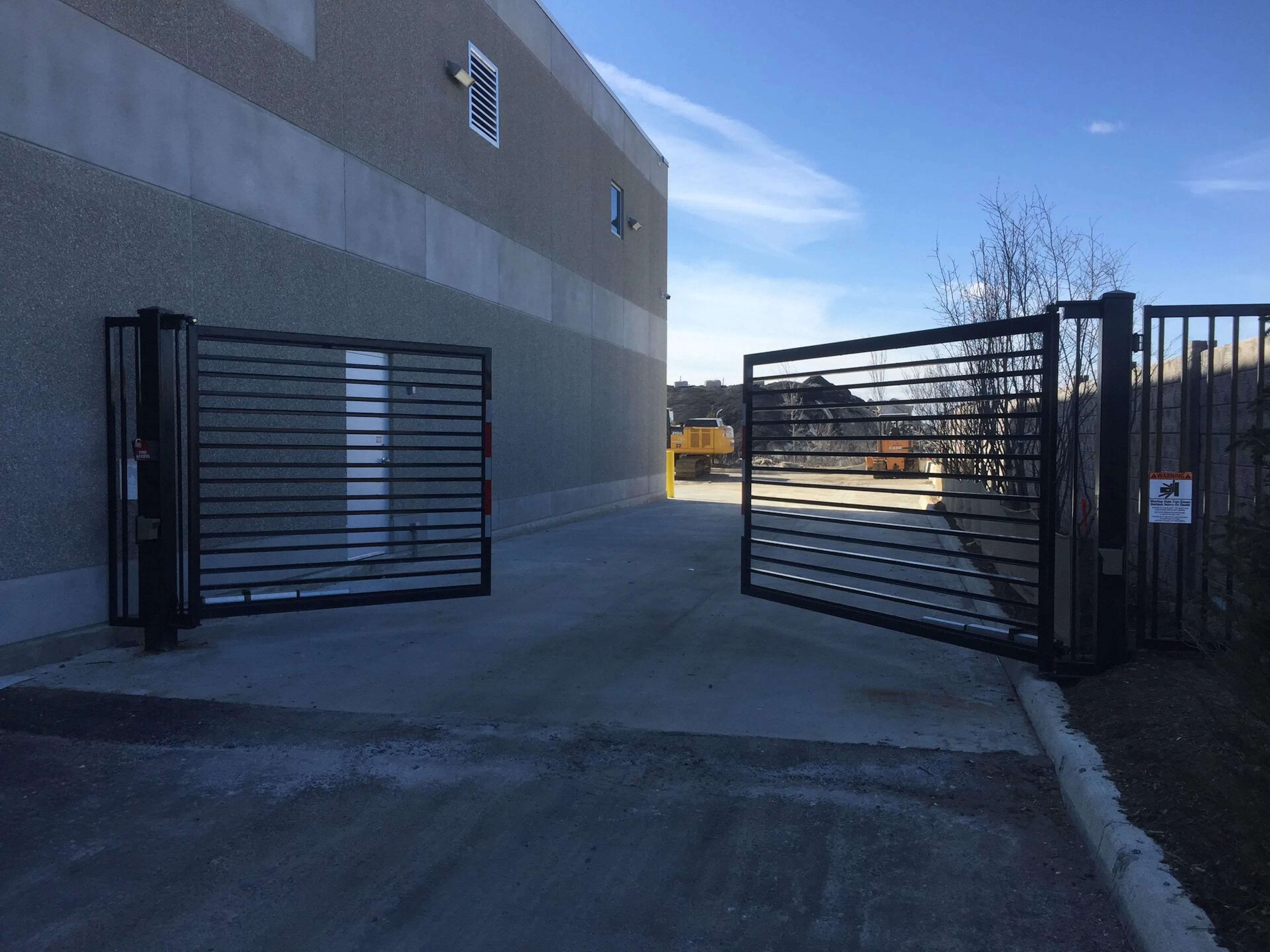 Swing metal security gates leading to a paved area beside a building, with clear blue sky in the background.