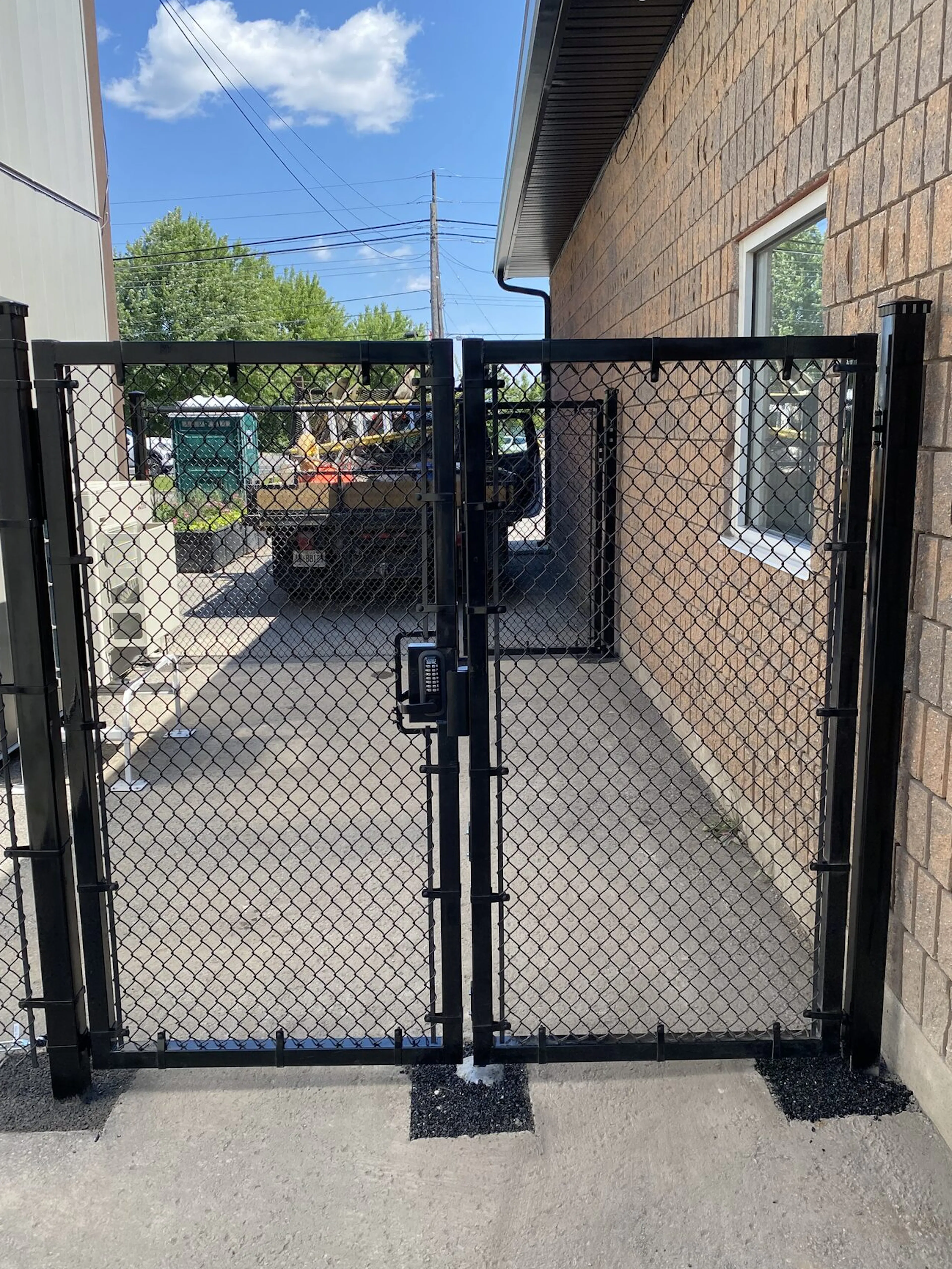 A black manual chain-link gate is installed between two fence posts leading to a paved area beside a building. The gate is revealing a concrete pathway and a truck visible in the background under a blue sky.