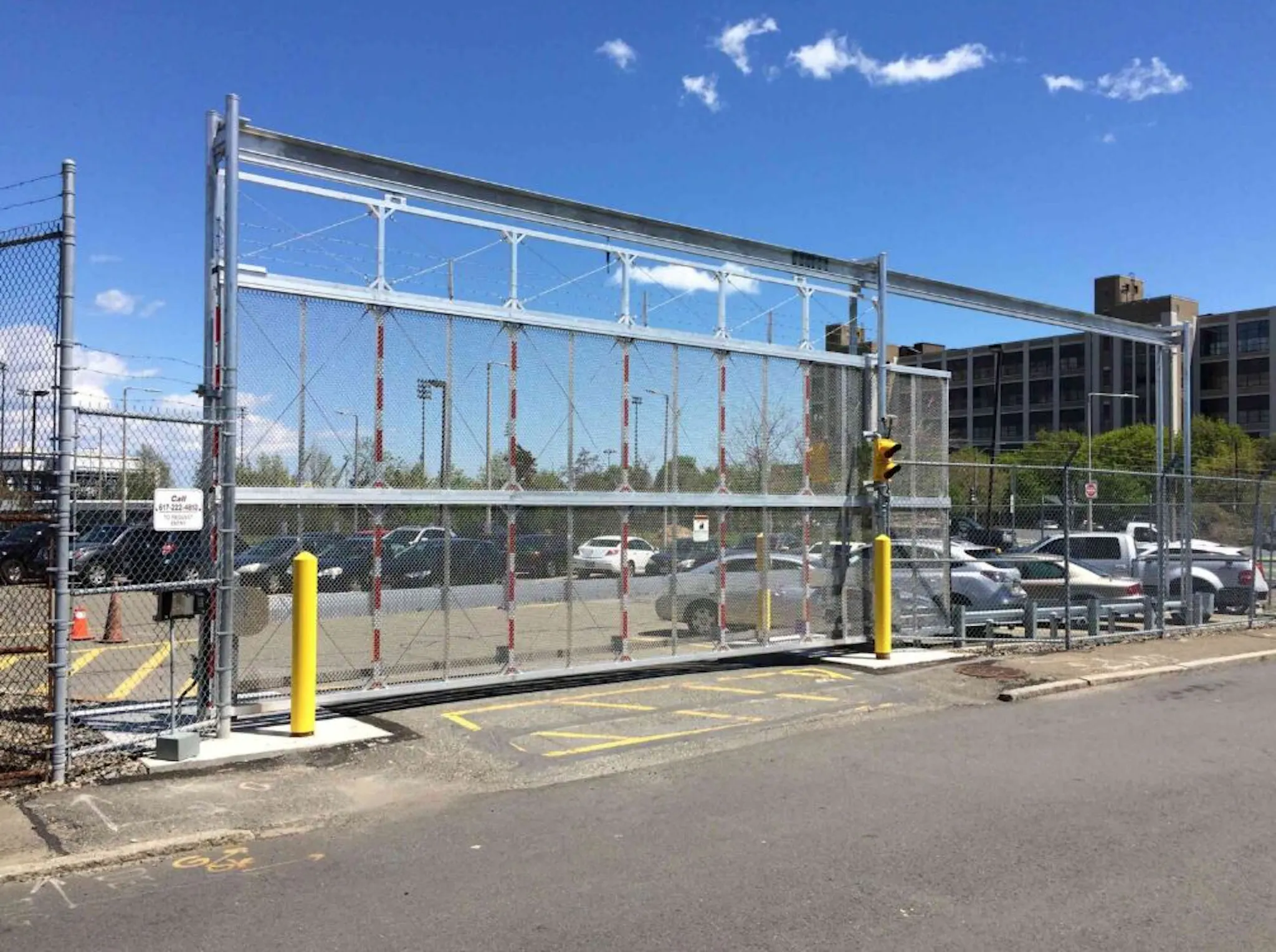 A large overhead metal security gate with a chain-link fence, yellow bollards, and a parking lot filled with cars under a clear blue sky.