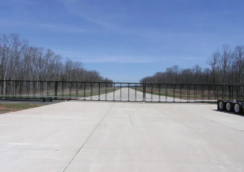 A large black oversized gate blocks the entrance to a wide road, flanked by asphalt pavement. The scene is set against a clear blue sky with sparse trees lining both sides of the road.