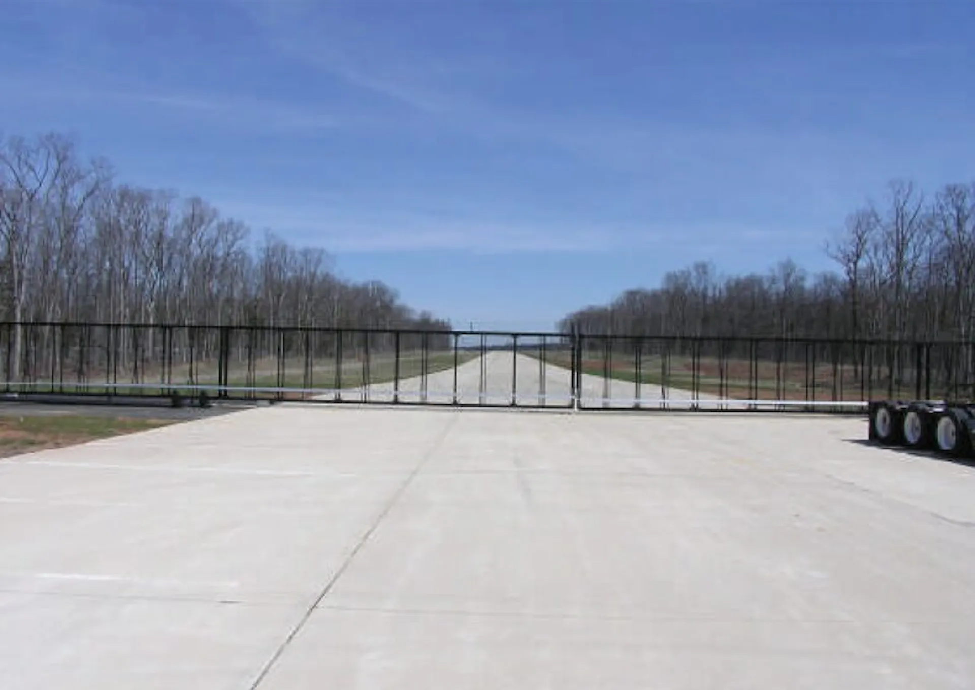 A large black oversized gate blocks the entrance to a wide road, flanked by asphalt pavement. The scene is set against a clear blue sky with sparse trees lining both sides of the road.