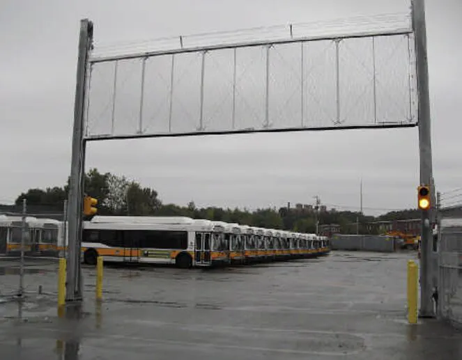 A line of buses parked in a transit terminal under a gray sky, with a grey chain-link vertical gate installed at the entrance. The area is wet, indicating recent rain, and there is a traffic signal visible near the entrance to the terminal.