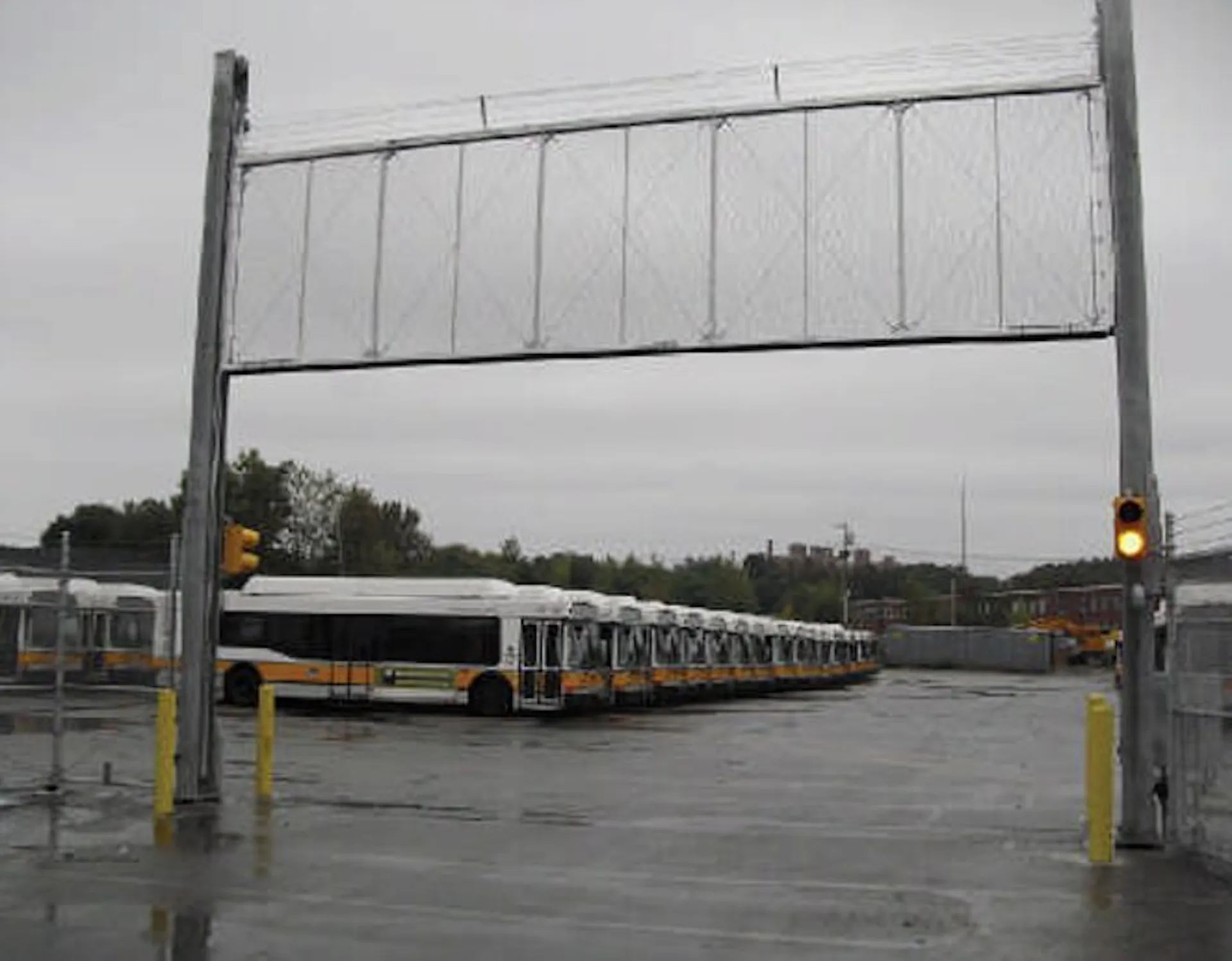 A line of buses parked in a transit terminal under a gray sky, with a grey chain-link vertical gate installed at the entrance. The area is wet, indicating recent rain, and there is a traffic signal visible near the entrance to the terminal.