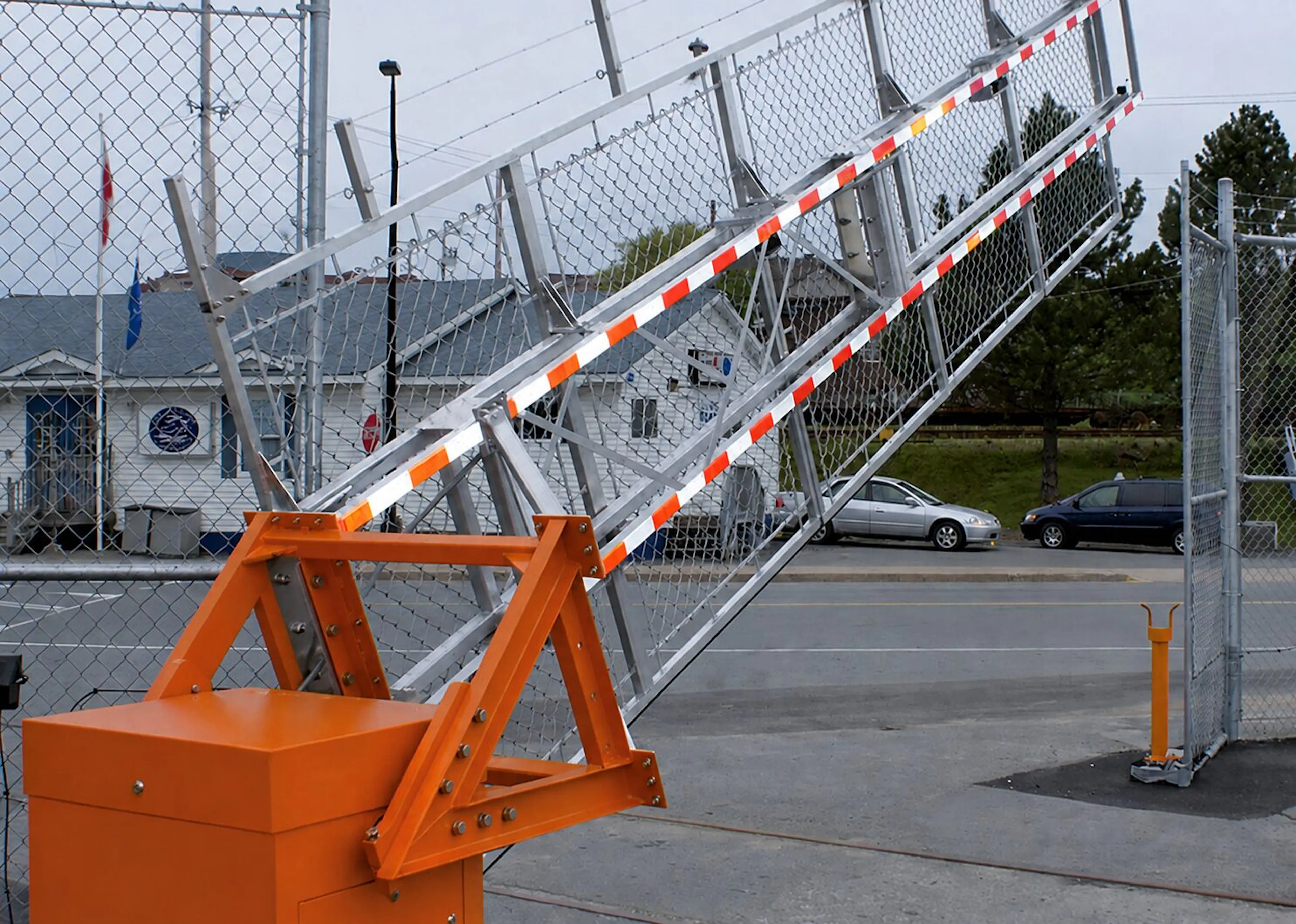 A vibrant orange barrier vertical pivot gate stands prominently at the foreground, its robust metal frame supporting a raised arm with alternating white and red stripes. The backdrop features a chain-link fence, leading into a spacious area with a few parked vehicles and a building visible in the distance, all under a cloudy gray sky.