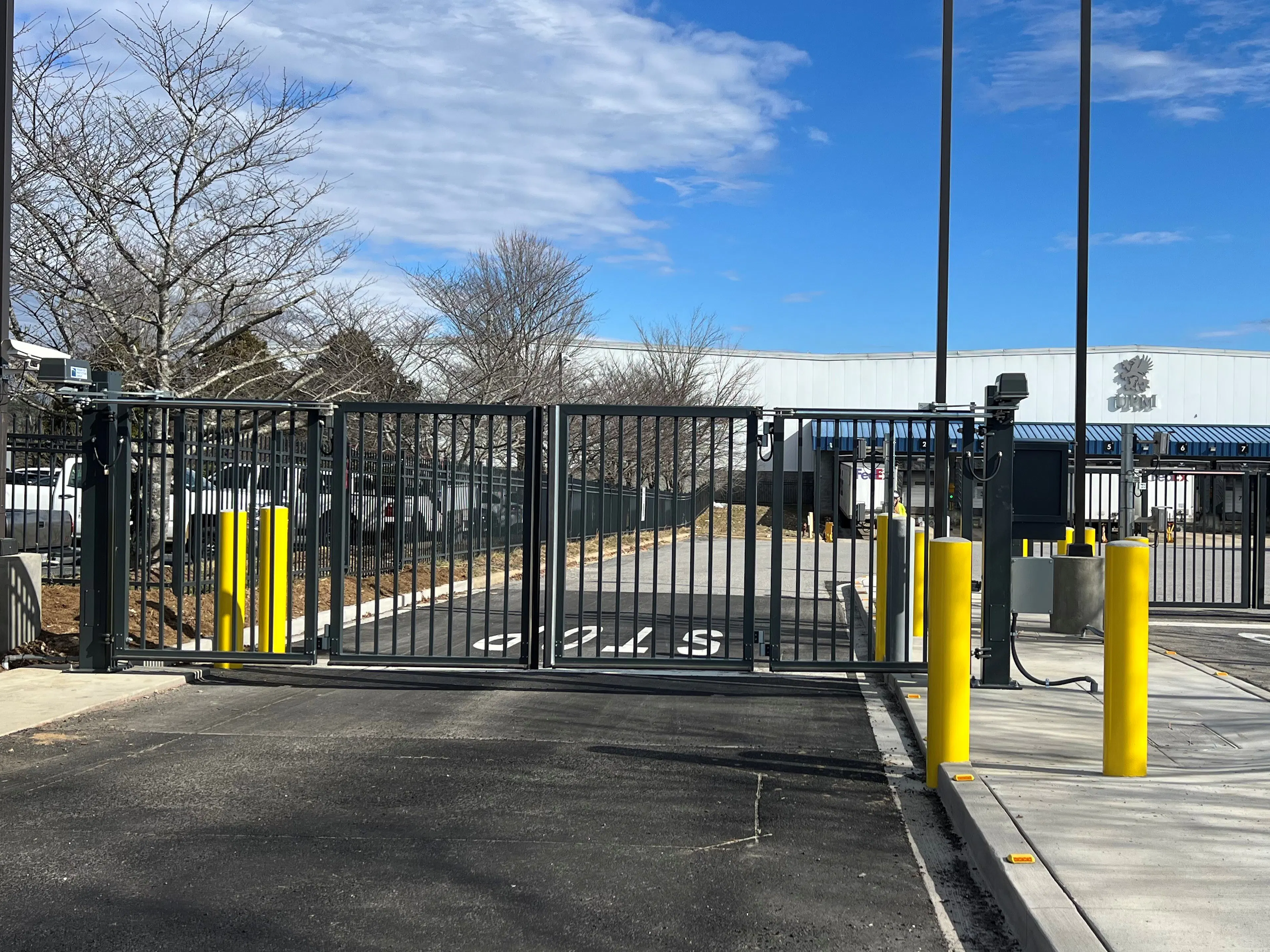 A black metal bi-fold gate, leading into a parking lot with asphalt. Yellow bollards are positioned near the gate entrance, and the background features gray trees and a white building under a blue sky with scattered clouds.