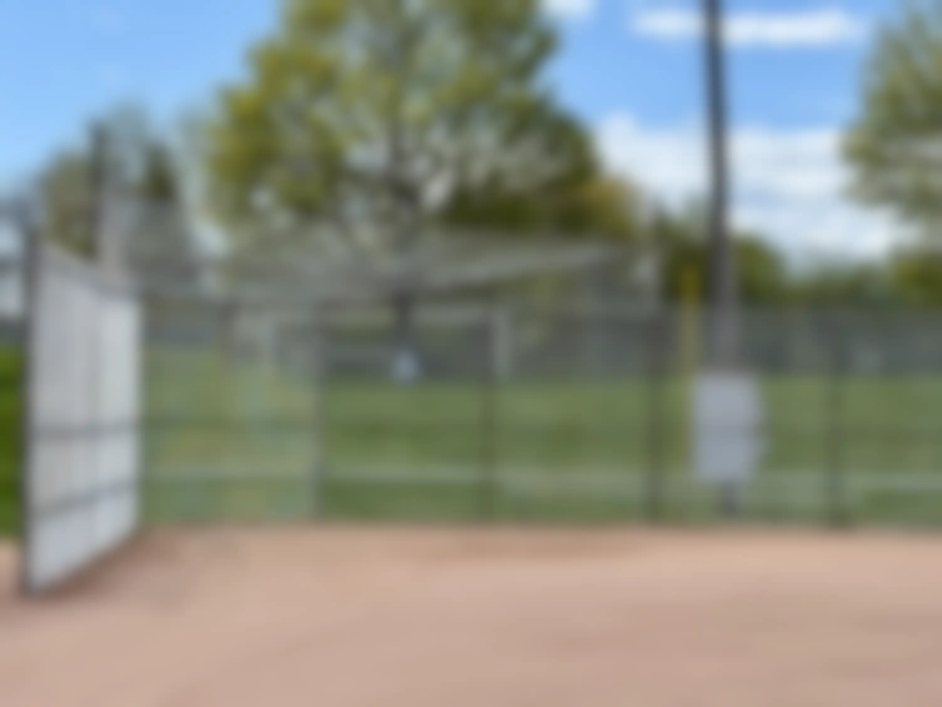 A gray chain-link fence surrounds a baseball field with a dirt infield and grass outfield.