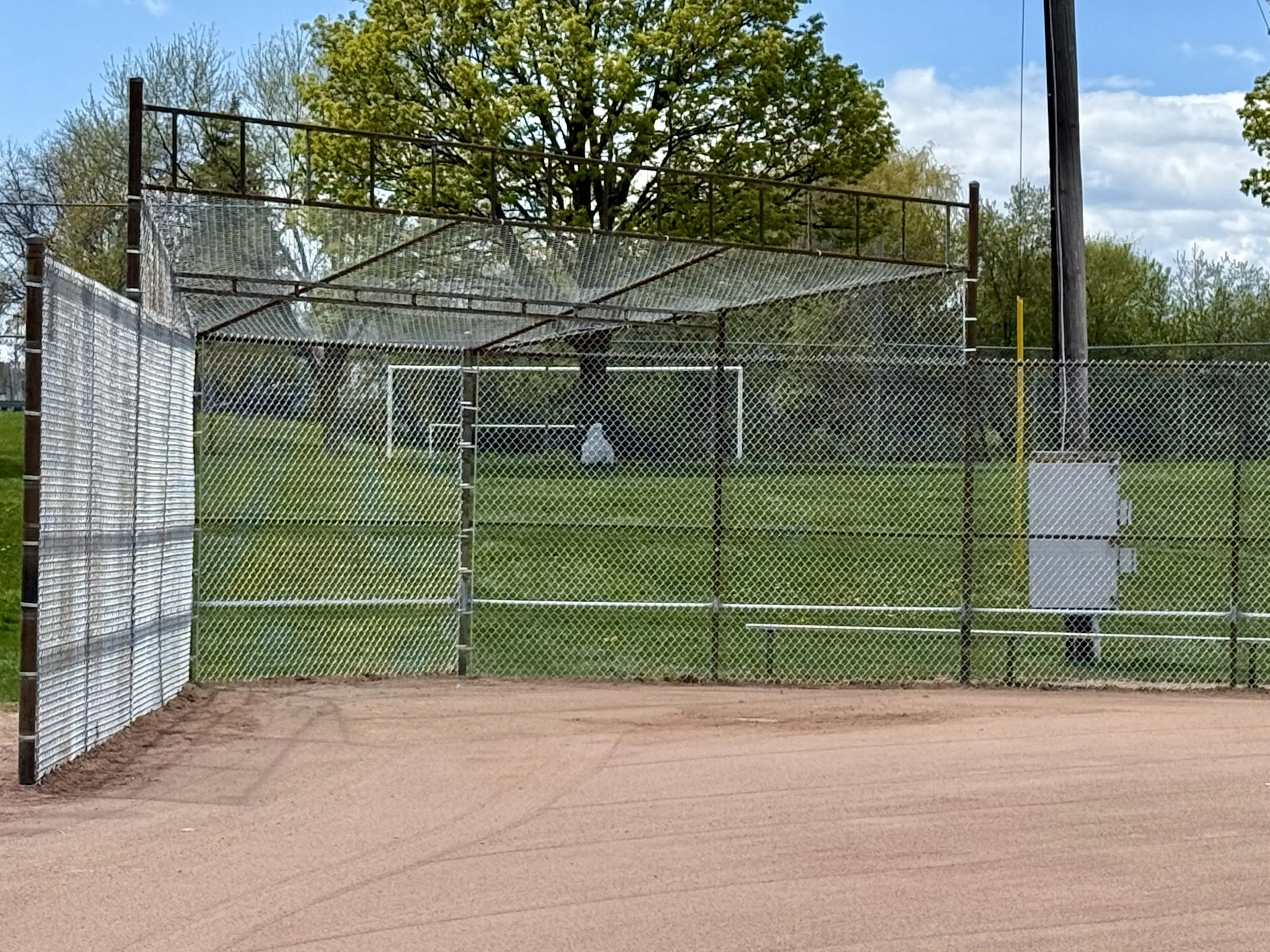 A gray chain-link fence surrounds a baseball field with a dirt infield and grass outfield.