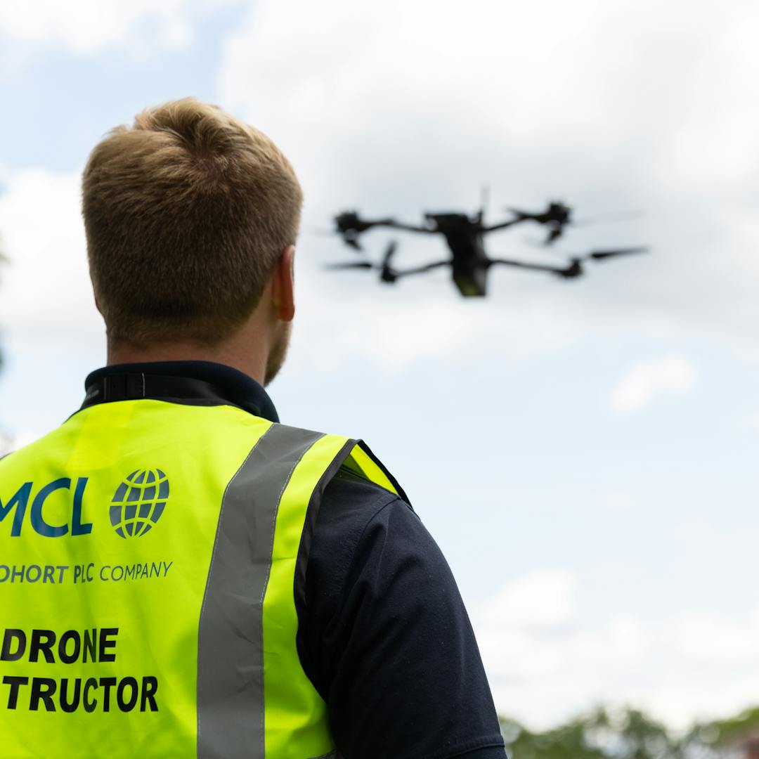 Drone instructor wearing high-visibility MCL safety vest observing a drone in flight against cloudy sky