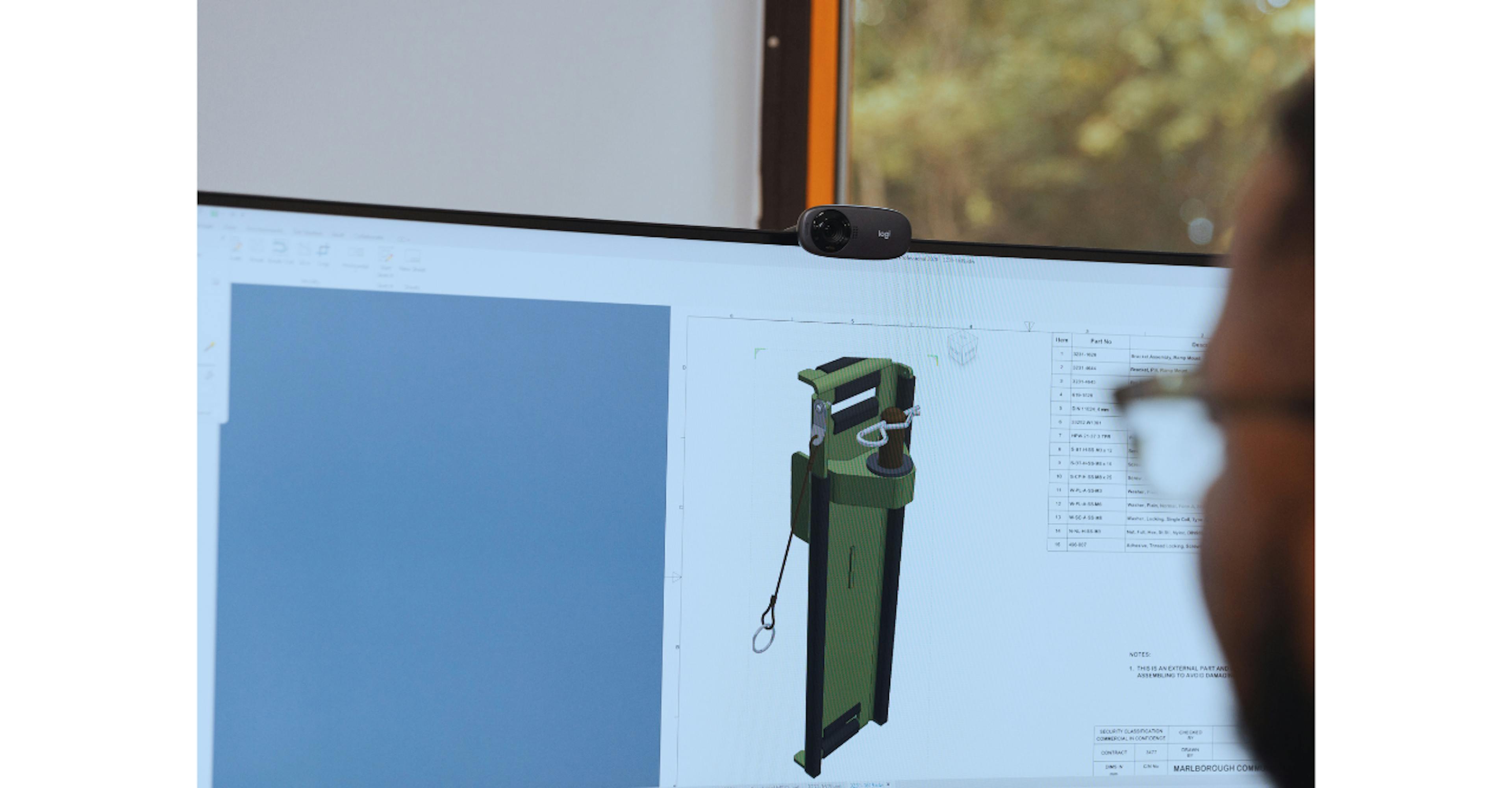 A person views a 3D CAD design of a green and black object on a computer monitor, with a webcam on top and a window with greenery in the background.