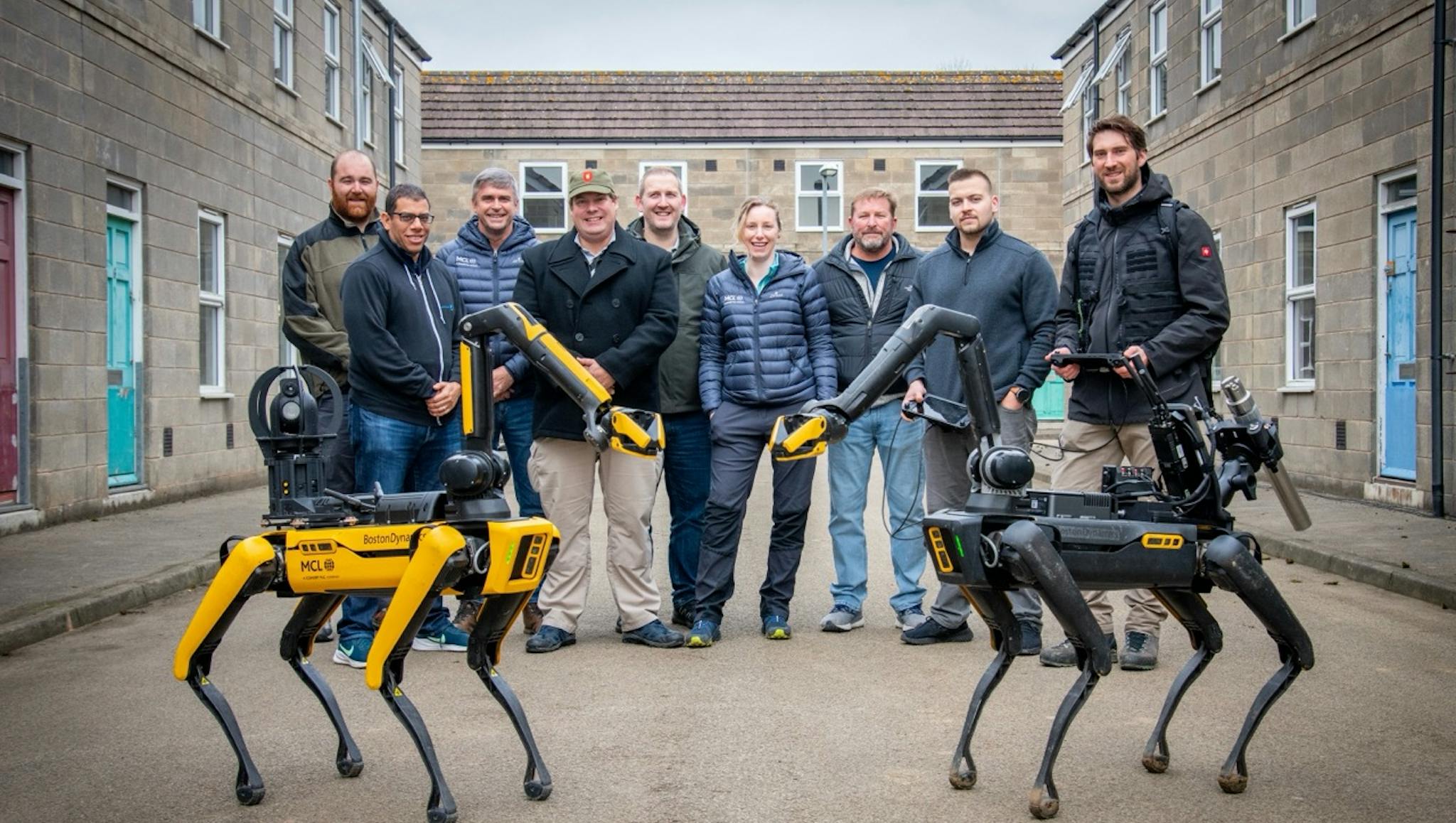 A group of nine people standing between two quadruped robots (Spot by Boston Dynamics) in a courtyard. The robots are equipped with specialised bomb disposal technology equipment - one yellow and one black. The group is positioned between concrete buildings with blue doors and windows. They are wearing casual and business casual attire including puffy jackets and sweaters. The yellow robot displays Marlborough Communications Ltd branding. 