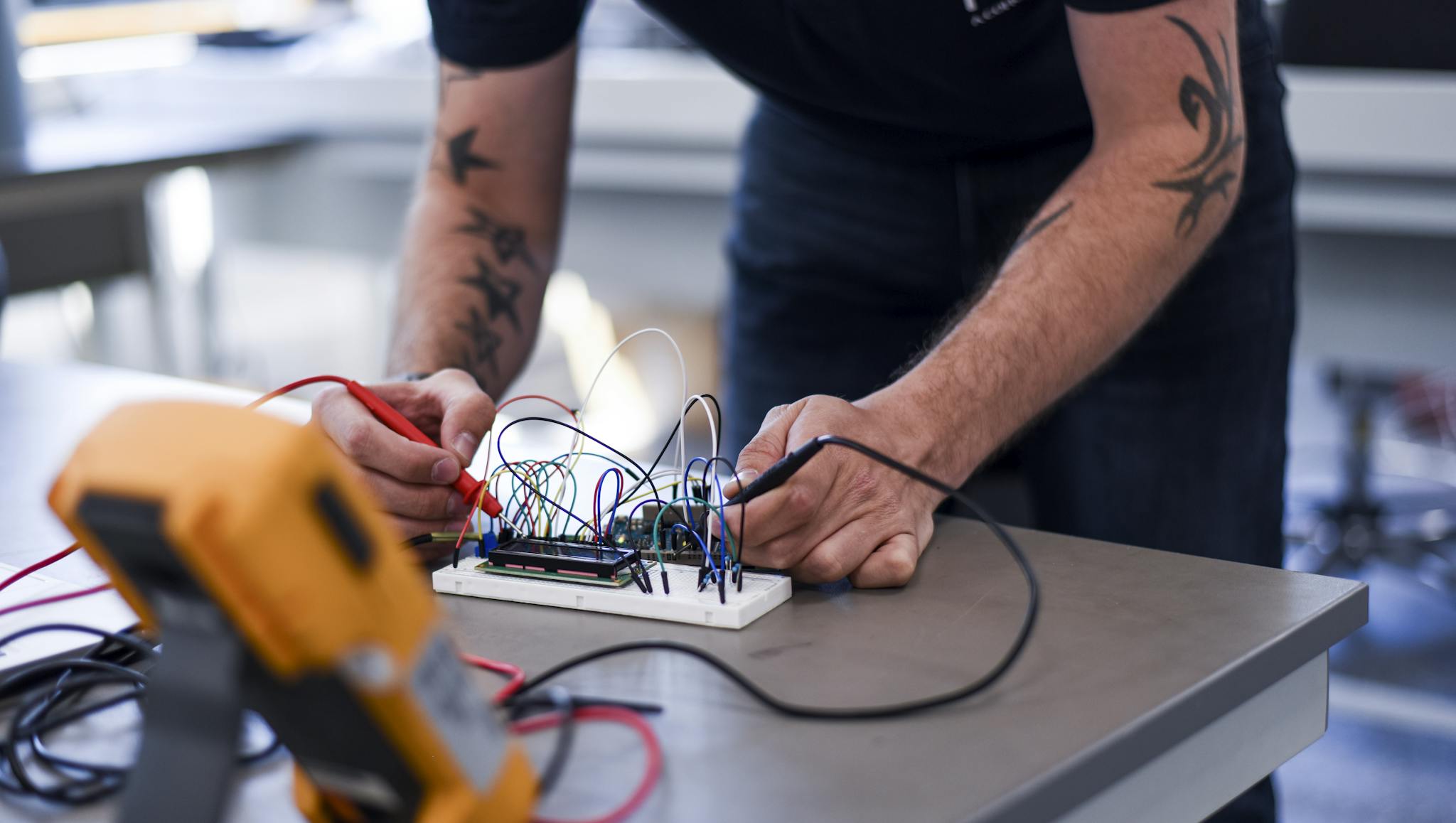 Engineer in an MCL shirt working on an electronic prototype circuit board, connecting wires with testing equipment visible on a workbench. Close-up view of hands manipulating electrical components during the development process