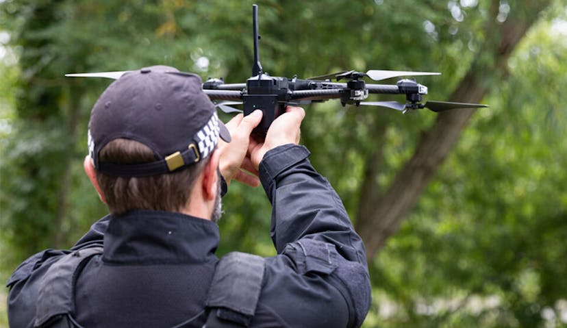 A police officer in tactical gear operates a drone outdoors, surrounded by greenery.