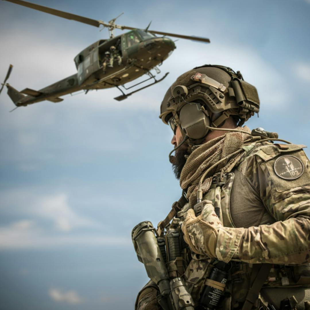 A soldier in camouflage gear and helmet looks on as a military helicopter flies nearby under a blue sky.