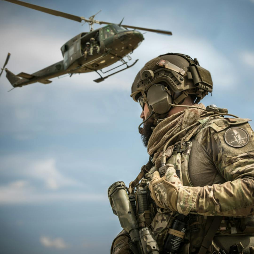 A soldier in camouflage gear and helmet looks on as a military helicopter flies nearby under a blue sky