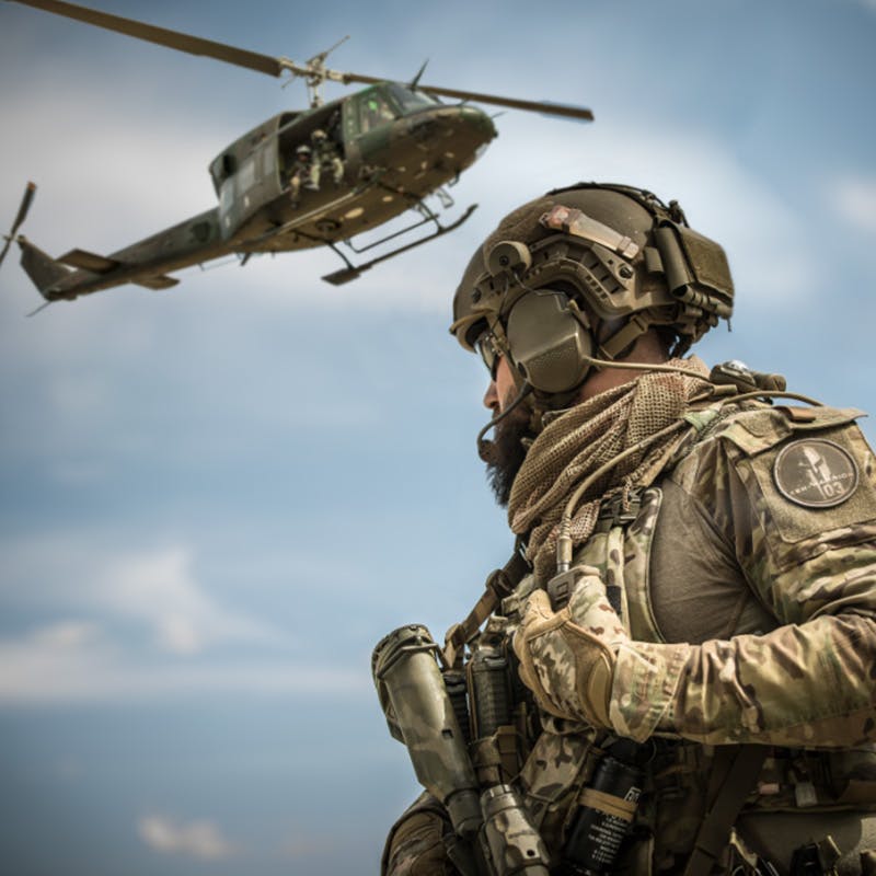A soldier in camouflage gear and helmet looks on as a military helicopter flies nearby under a blue sky