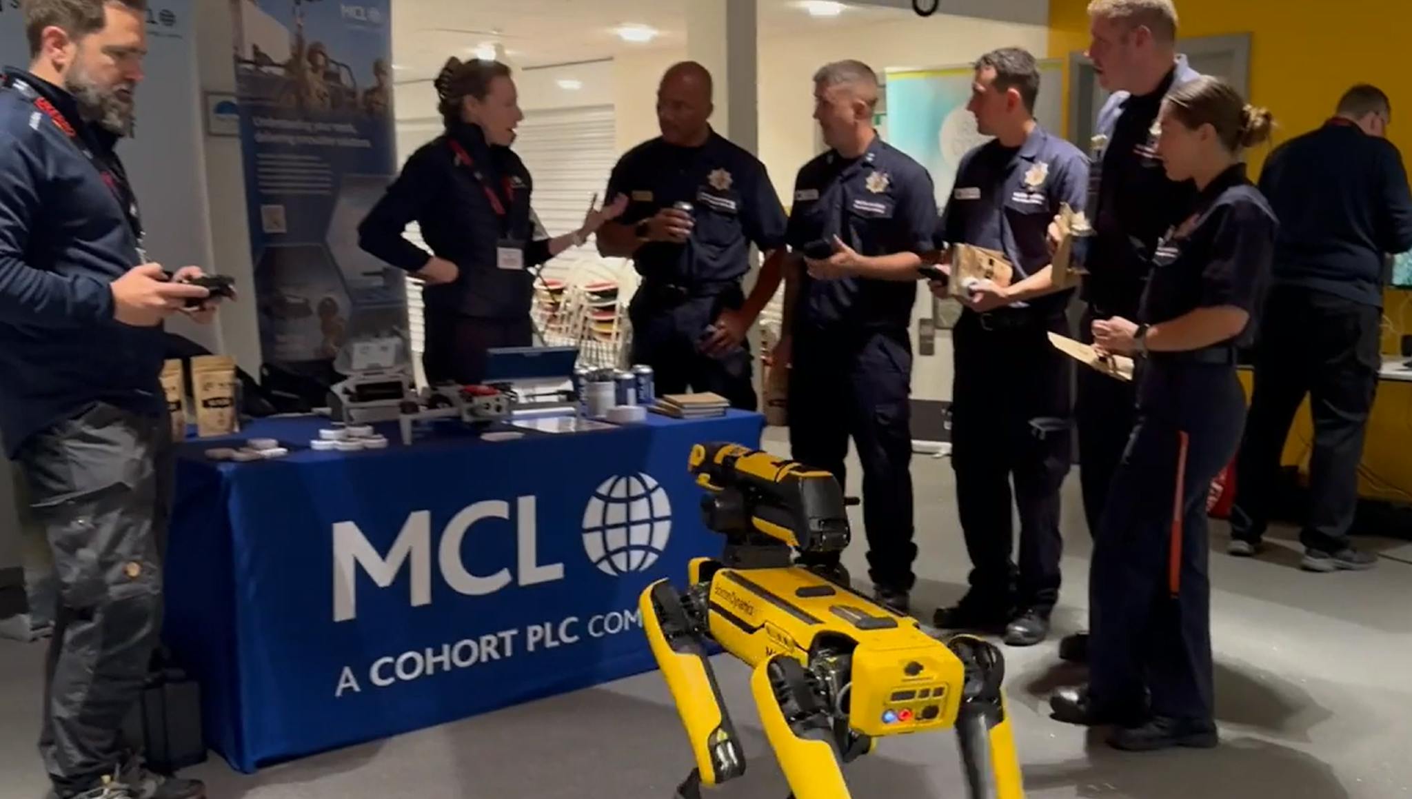 Group of uniformed emergency personnel gather around MCL company display with yellow quadruped robot (Spot) in foreground.