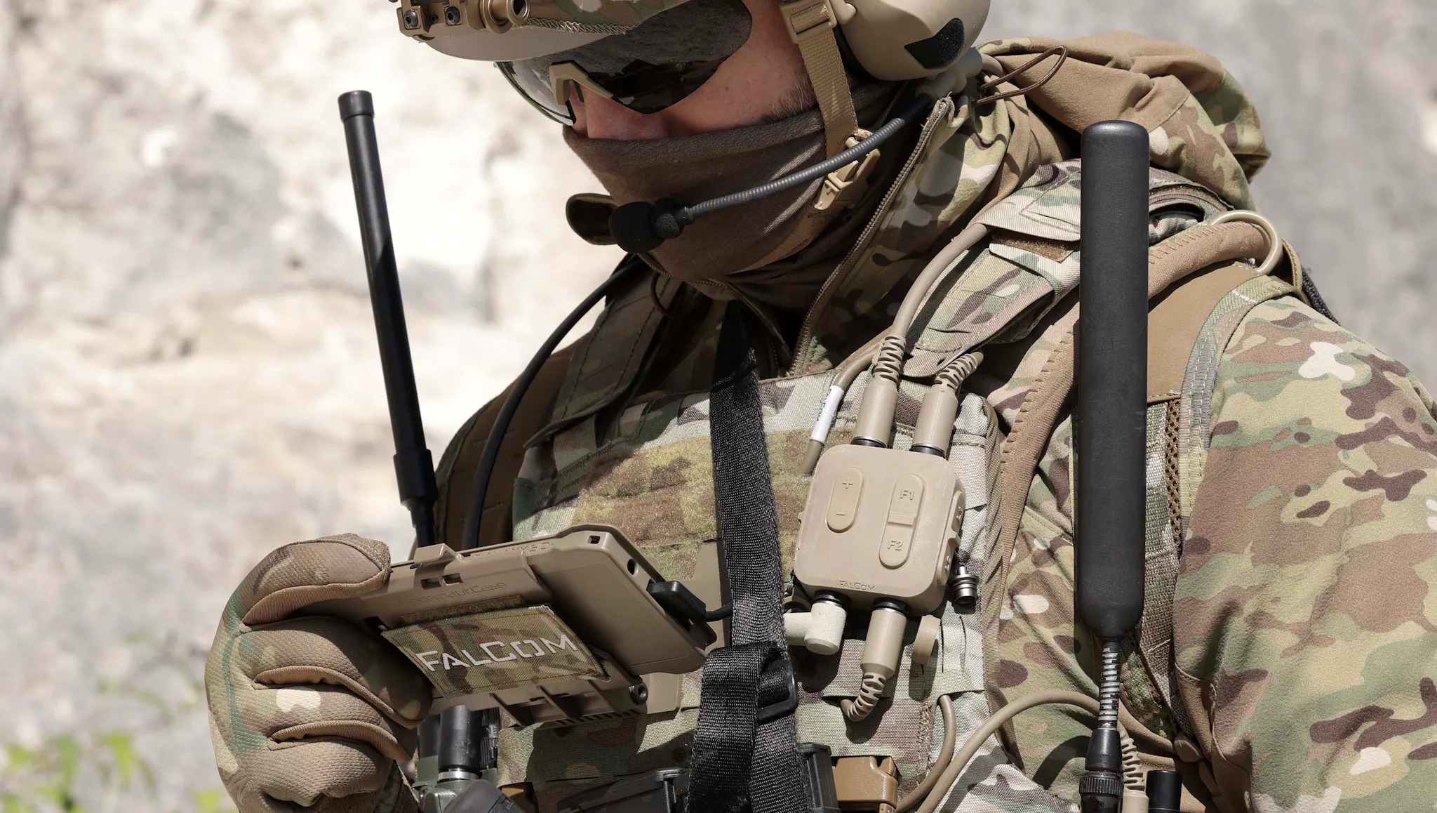 A soldier in full combat equipment, with a FalCom multi-function control unit and cabling visible on their chest rig