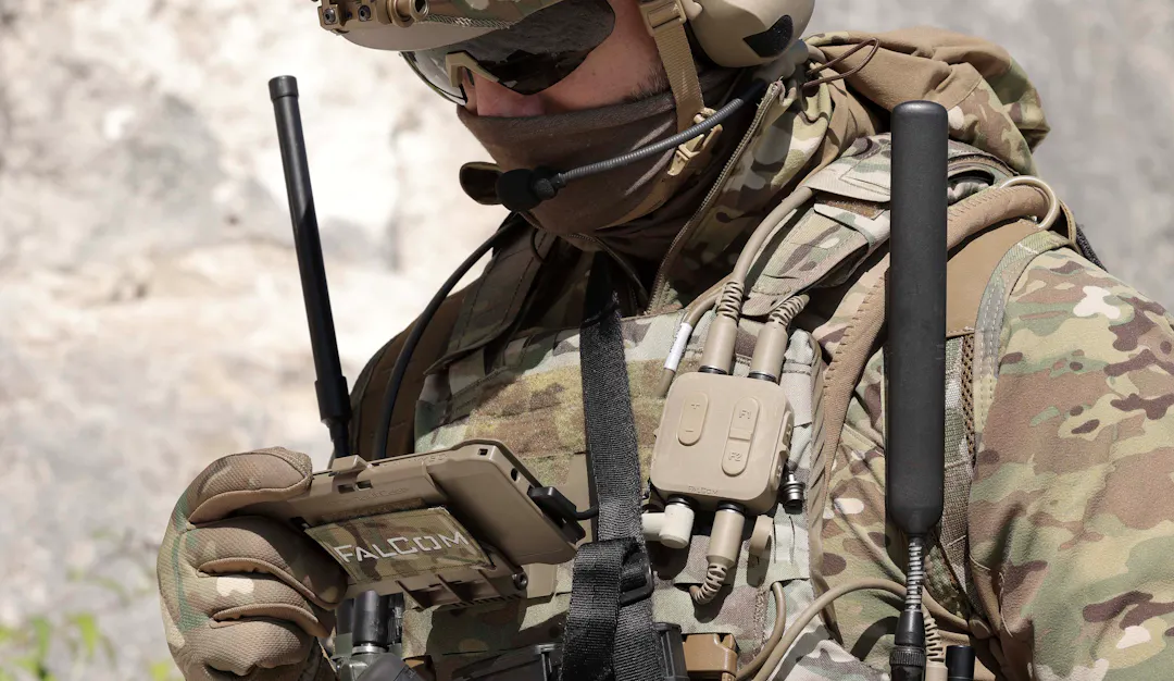 A soldier in full combat equipment, with a FalCom multi-function control unit and cabling visible on their chest rig