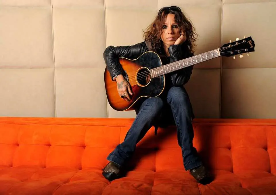 Linda Perry sat on back of sofa with acoustic guitar